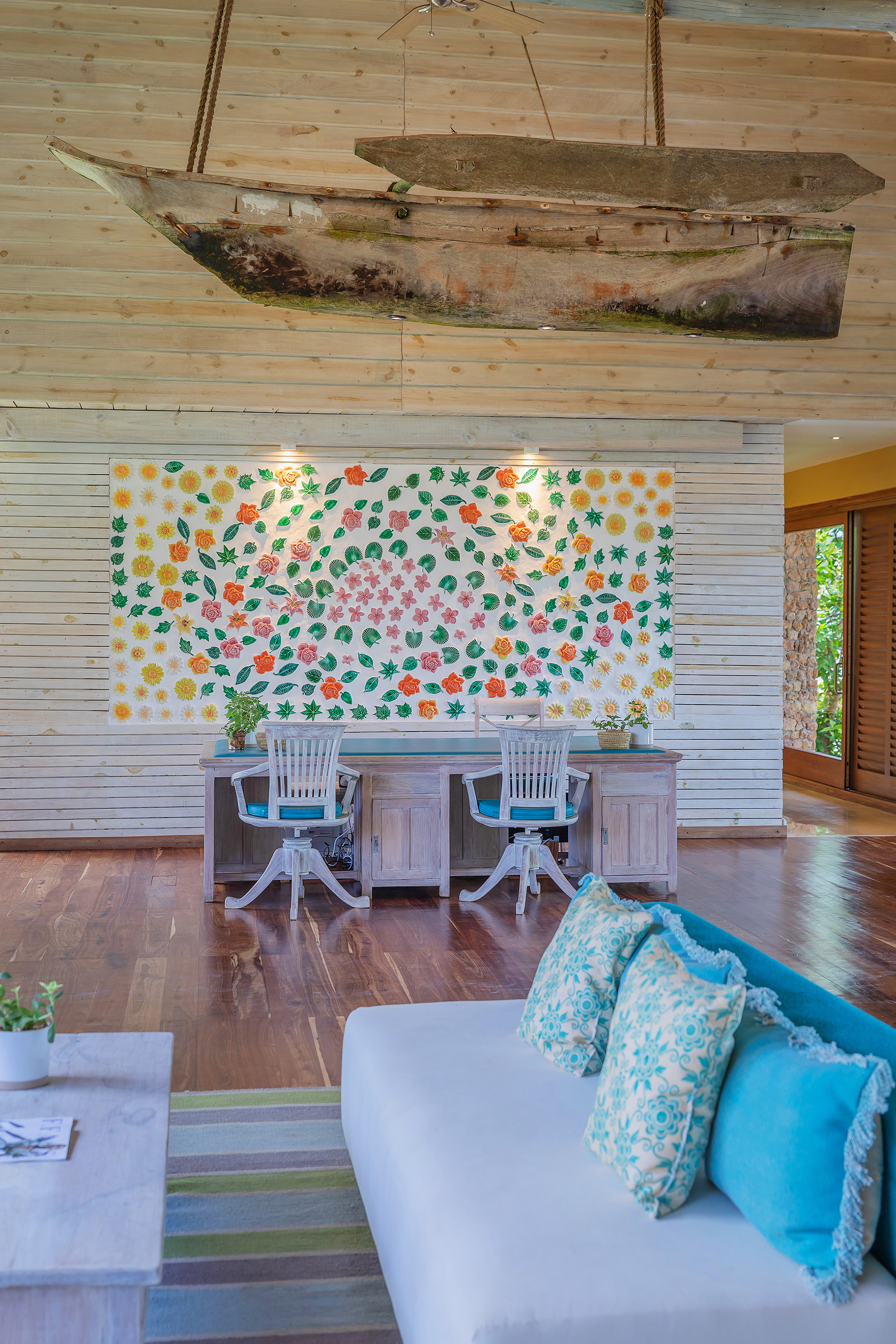 Tropical interior: hanging boat, floral mural, wooden desk, and white sofa.