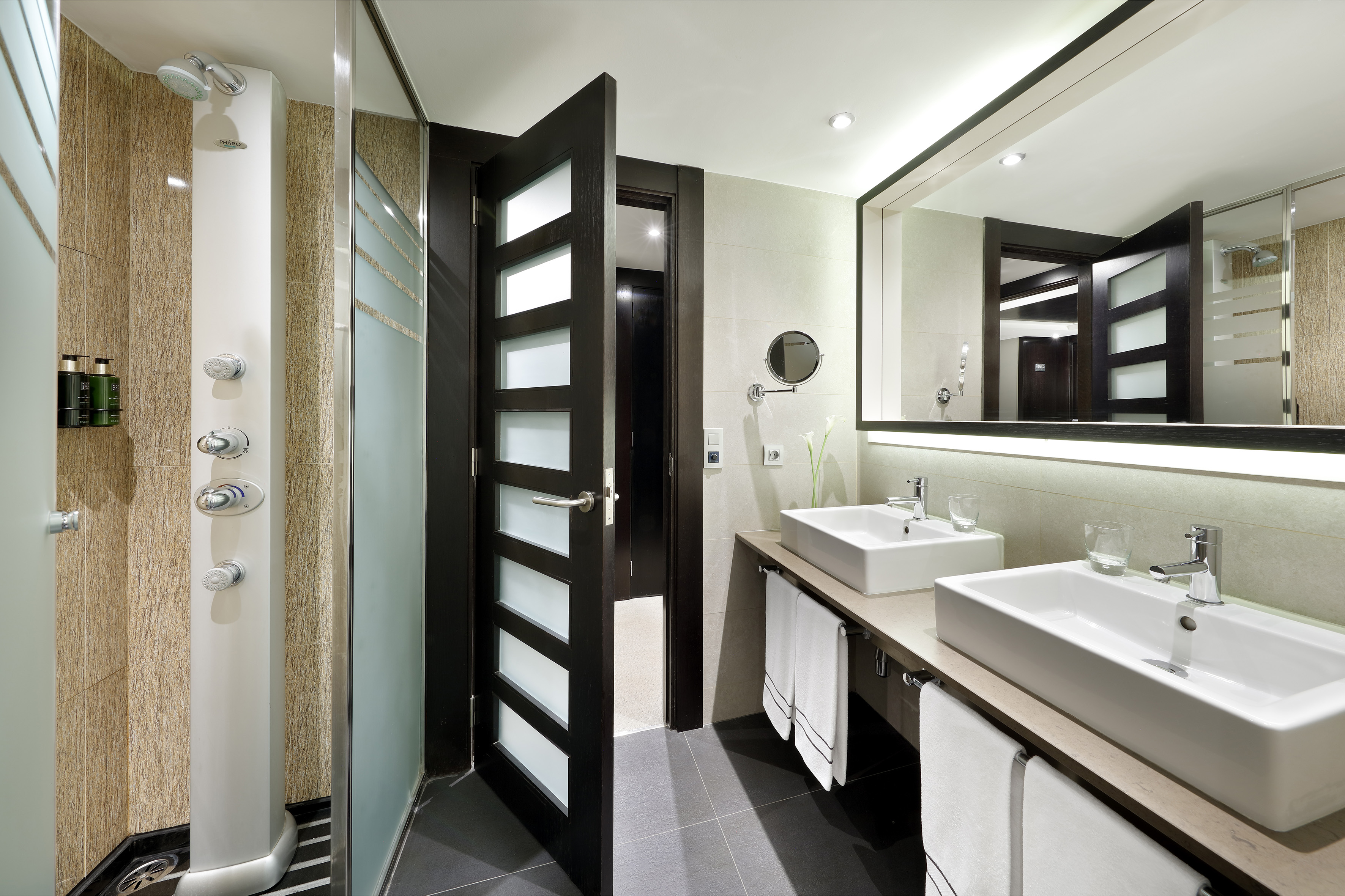 Modern hotel bathroom with walk-in shower, dual vanity, and backlit mirror.