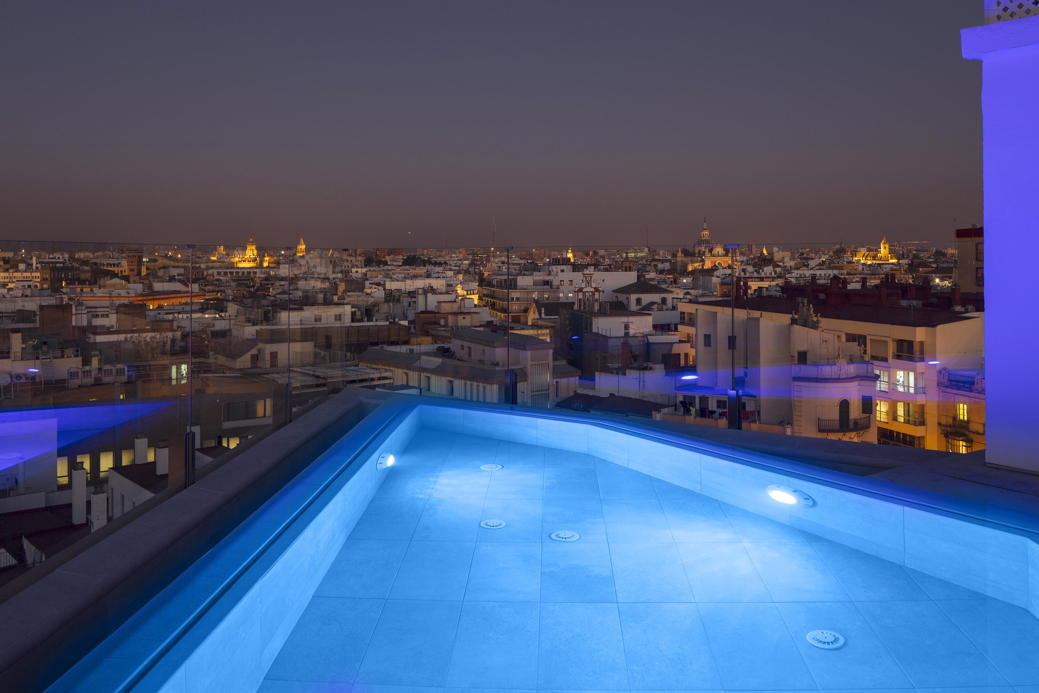 Blue lit rooftop jacuzzi with city skyline view at night.