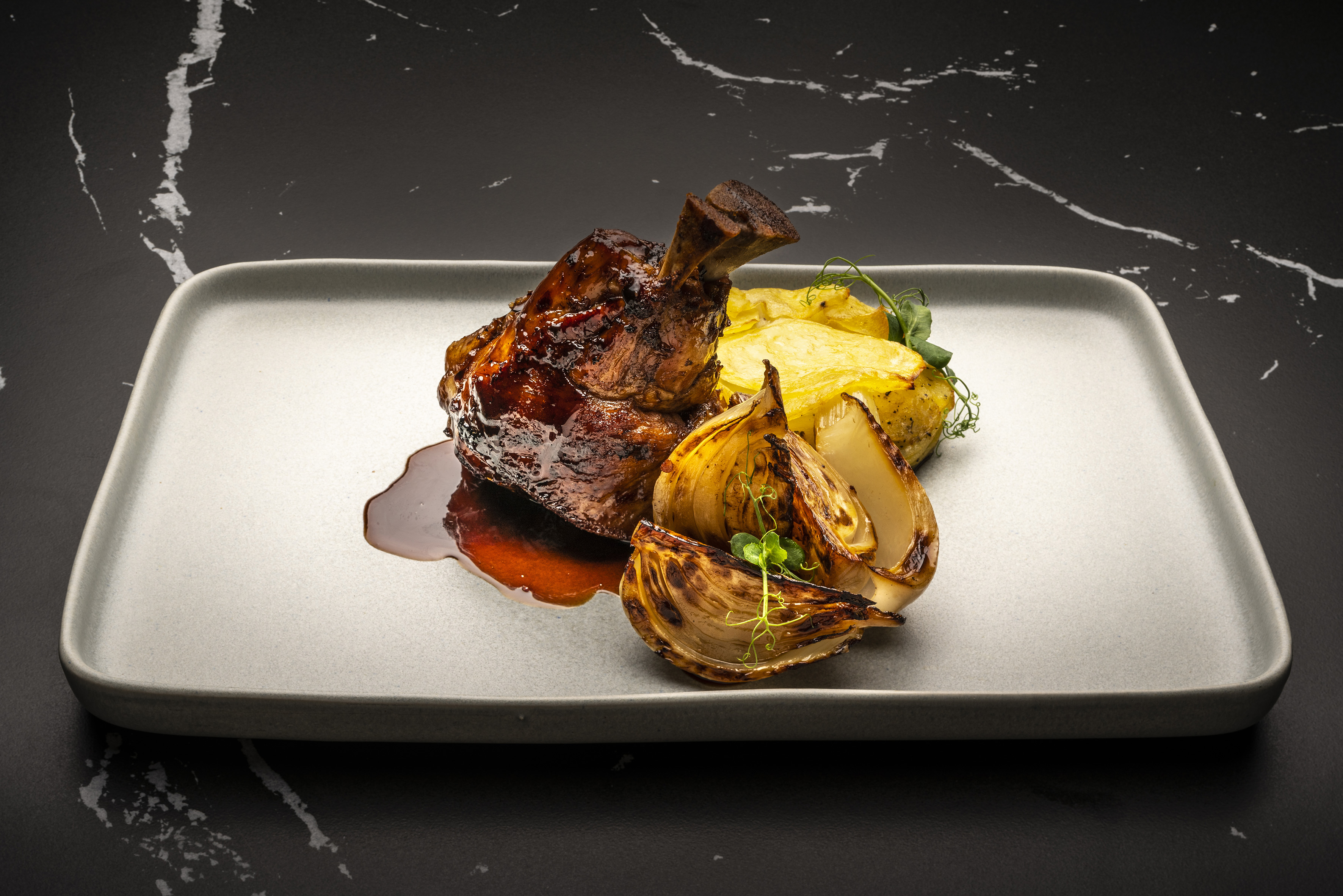 Glazed meat shank, roasted potatoes, and charred onions on a gray plate.