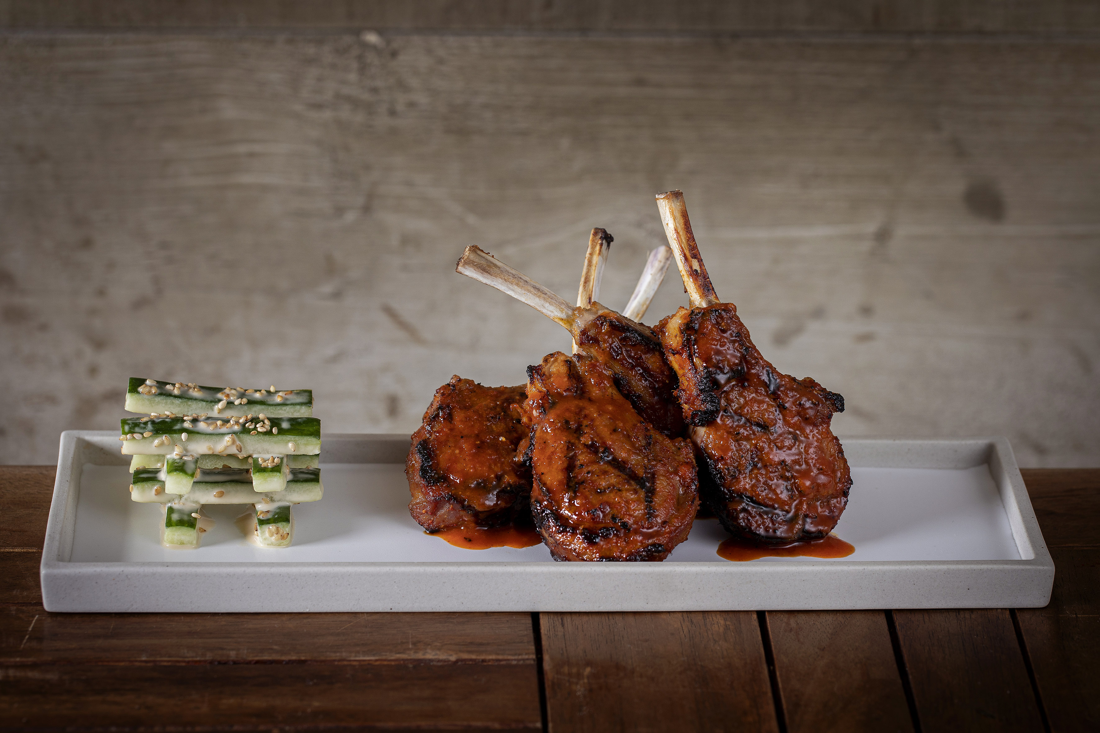 Grilled lamb chops with sauce and stacked cucumber sticks on a white plate.