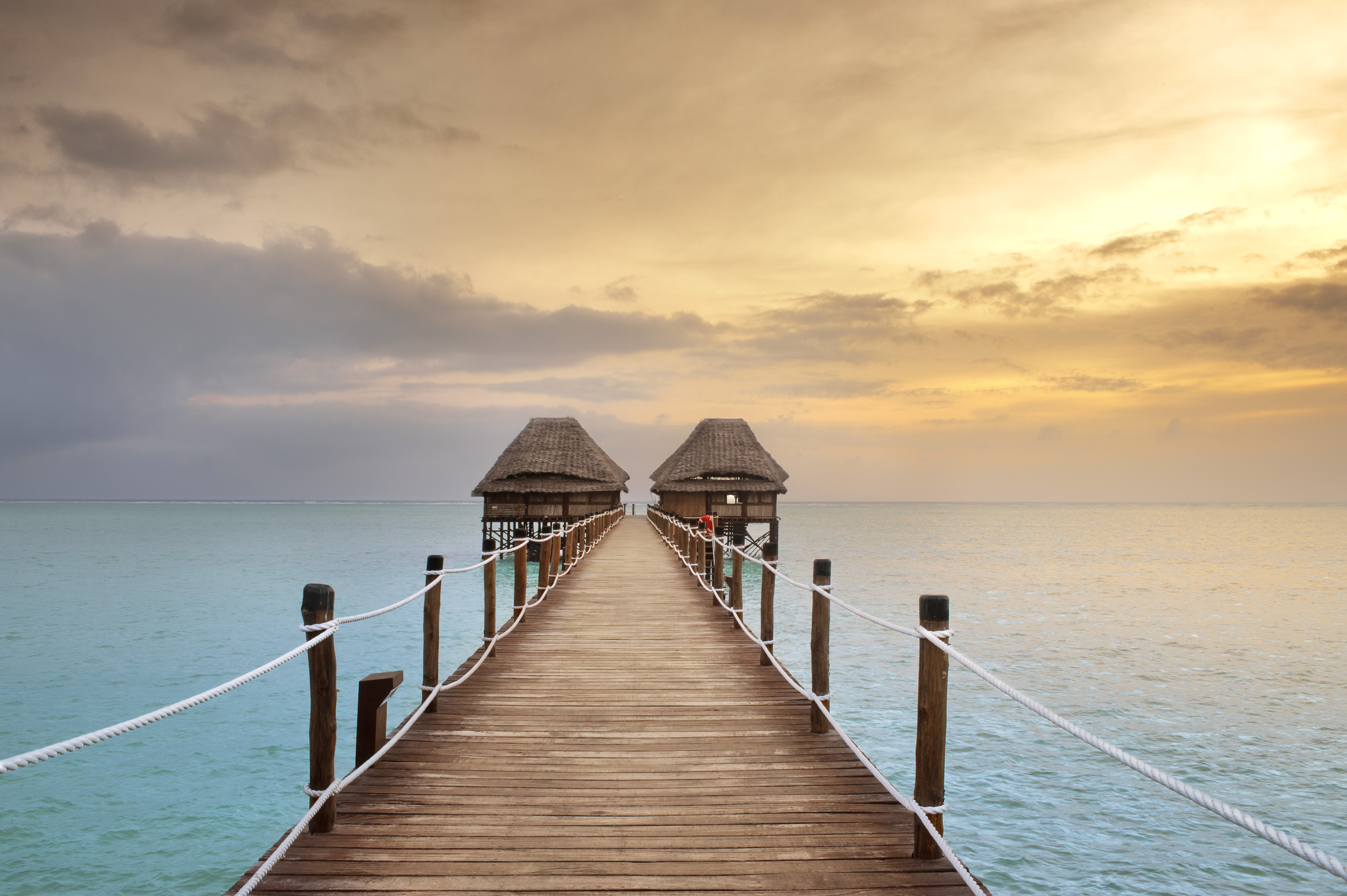 Tropical wooden pier to two overwater bungalows at sunset.