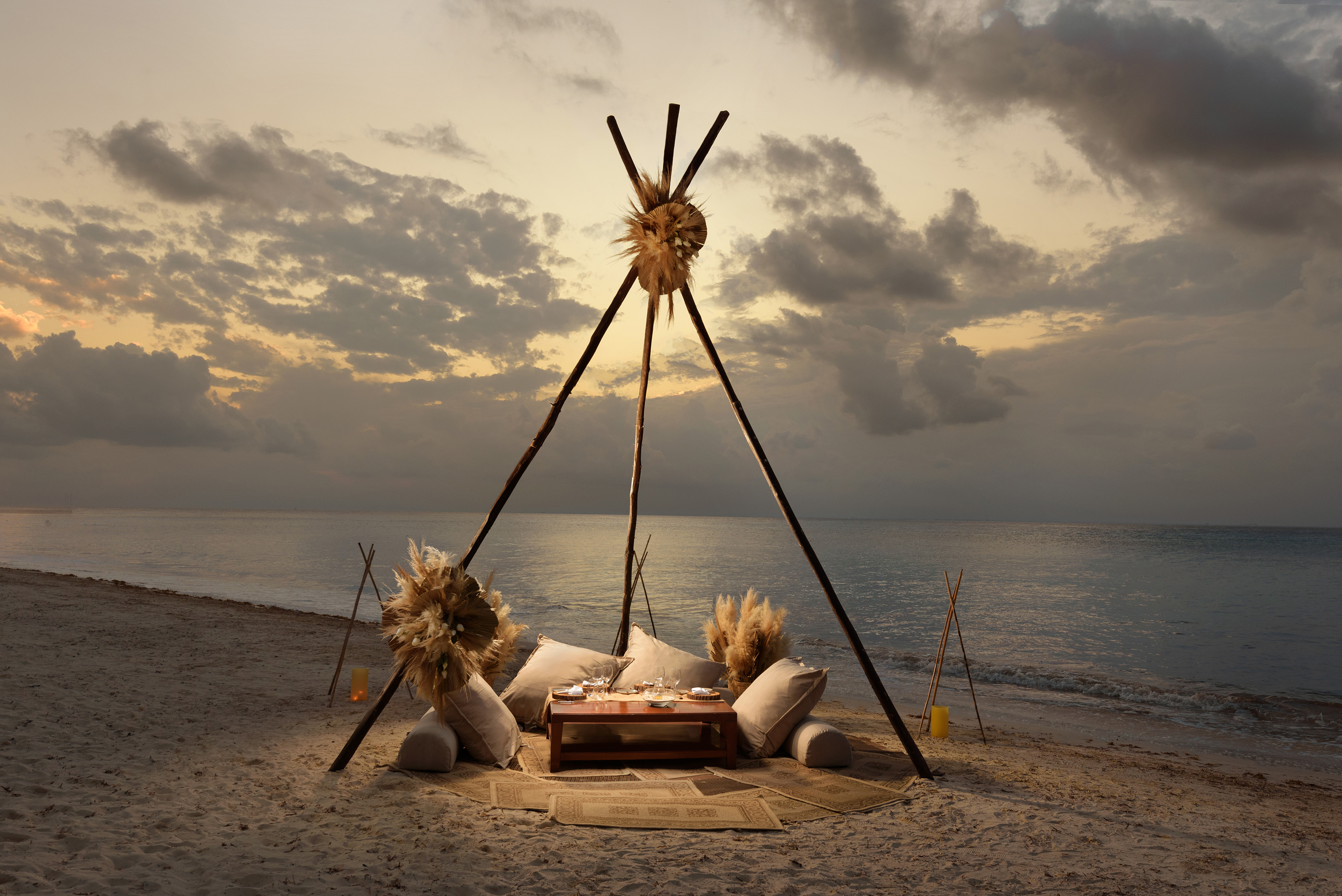 Sunset beach dining setup with teepee, cushions, and ocean view.
