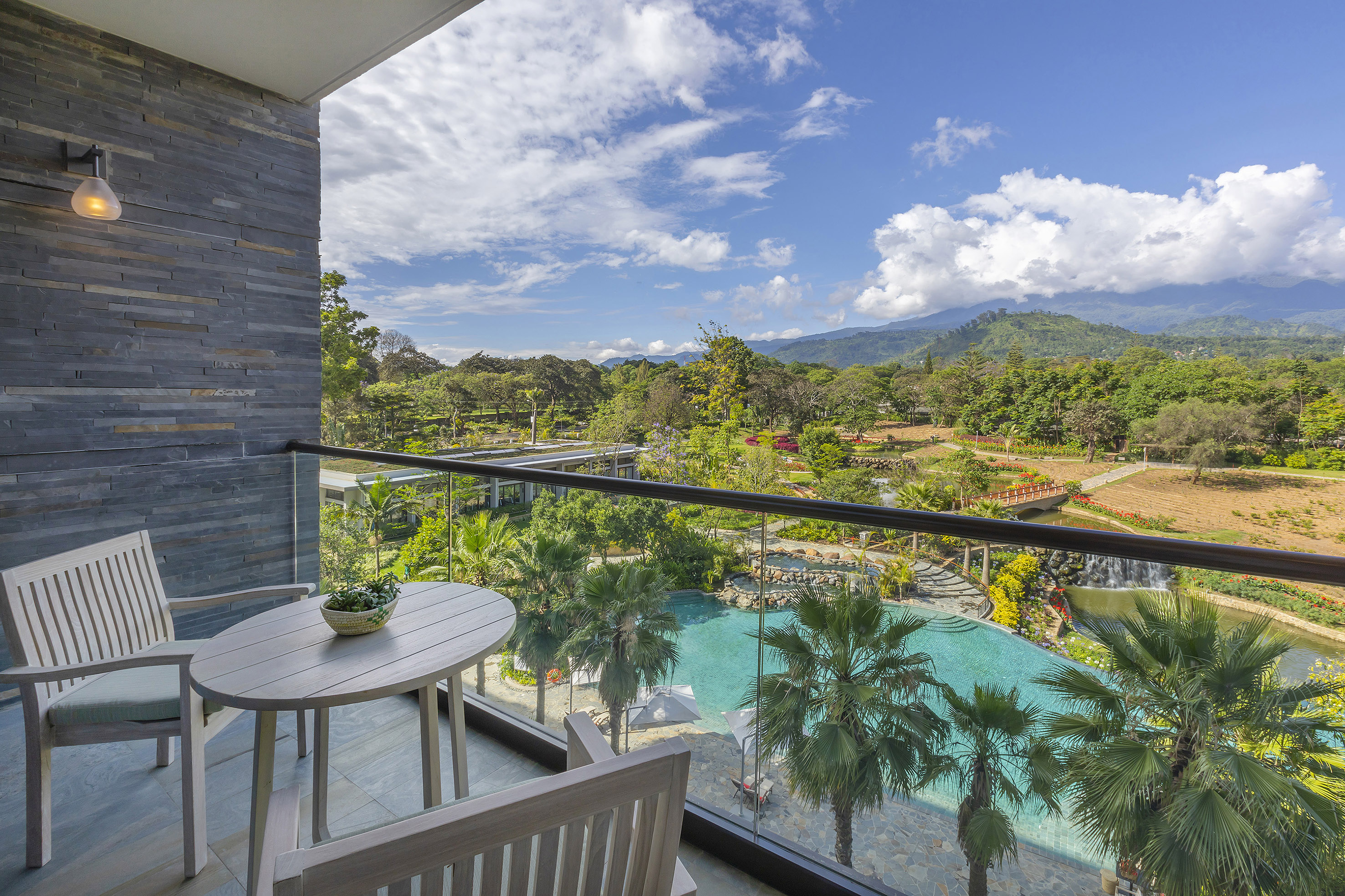 Resort balcony with outdoor seating, pool, palm trees, and scenic mountain views.