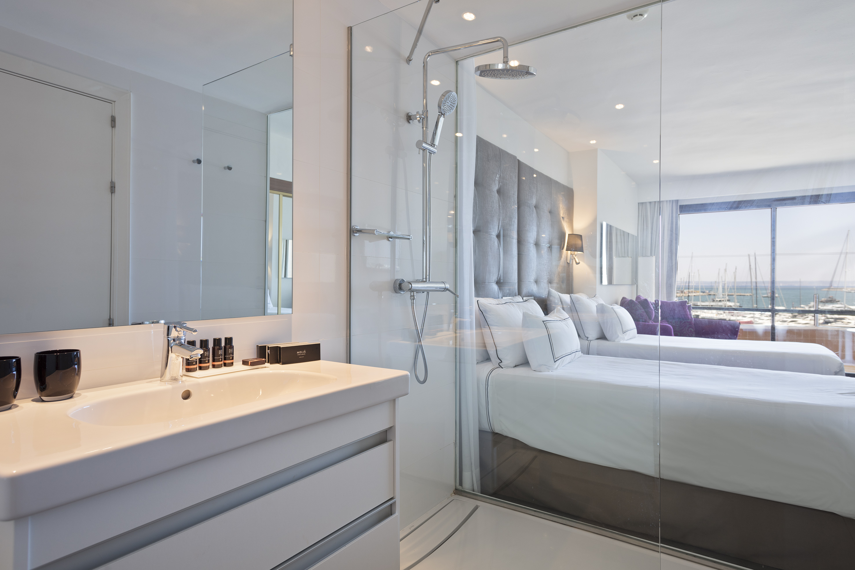 Modern hotel bathroom with glass shower, sink, and bedroom overlooking a marina.