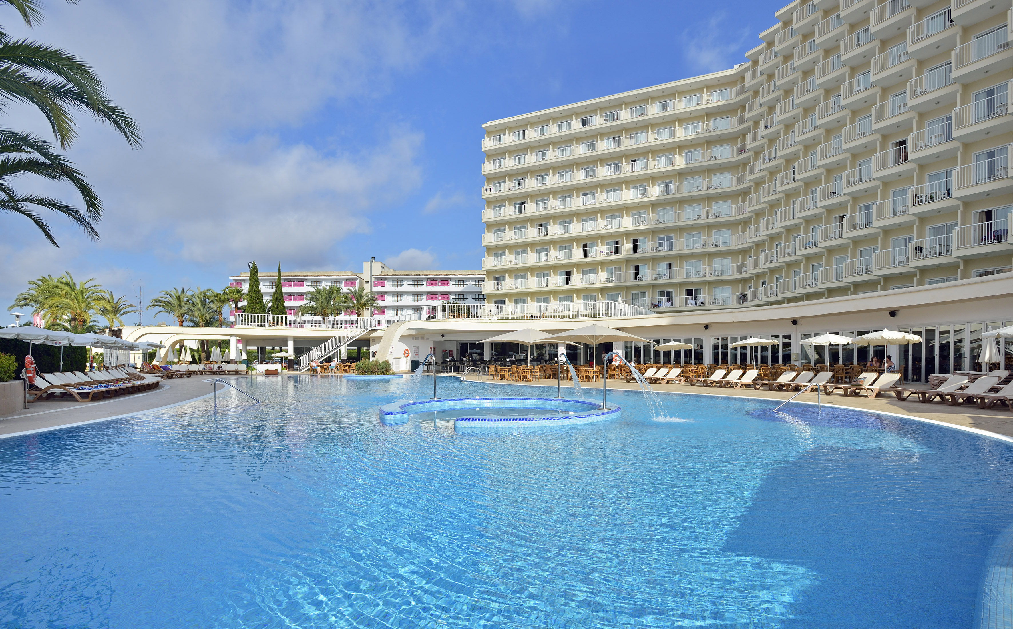 Expansive hotel resort with large outdoor pool, sun loungers, and palm trees.