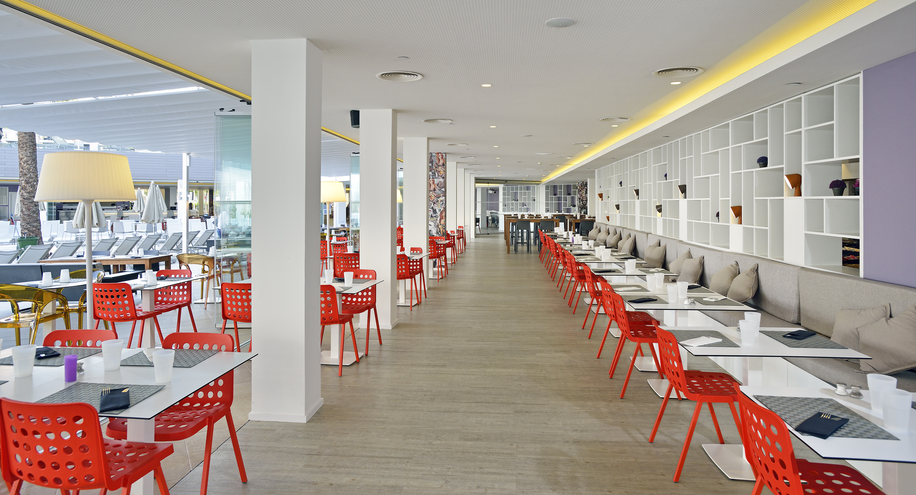 Bright modern dining room with red chairs, white tables, and poolside view.