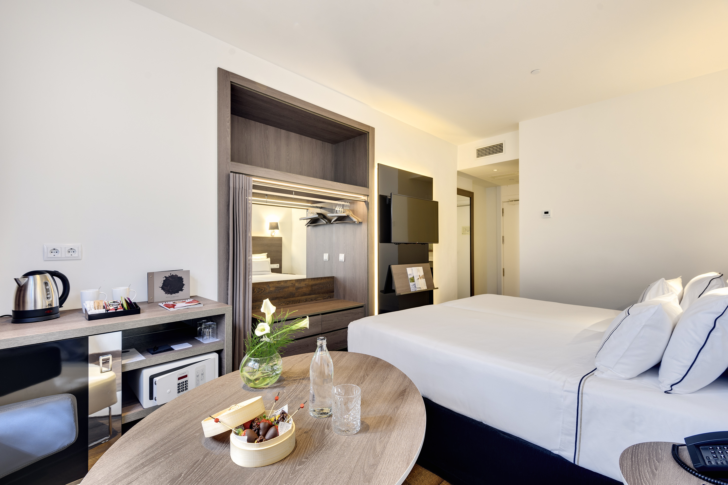 Modern hotel room with twin bed, desk, minibar, and fruit dessert.