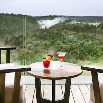 a table with a drink on it and chairs on a deck with a view of a waterfall