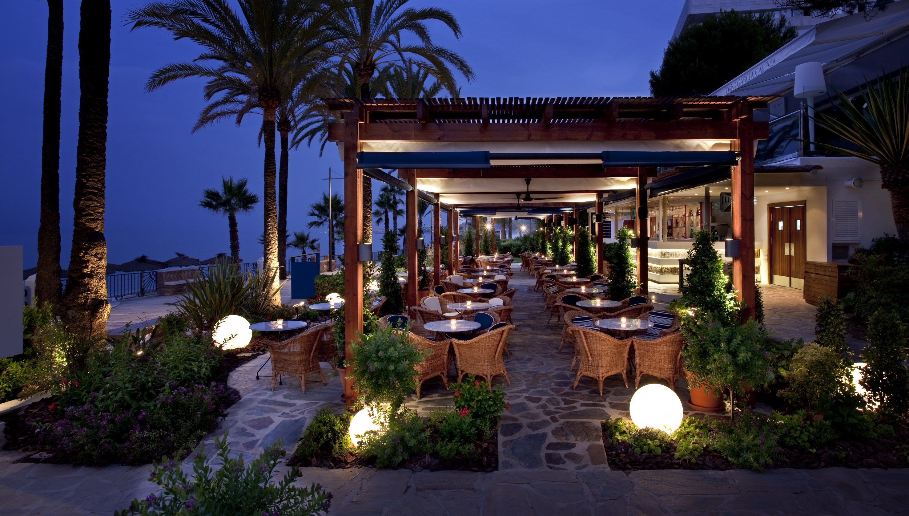 Illuminated outdoor restaurant patio with palm trees and wicker chairs at night.
