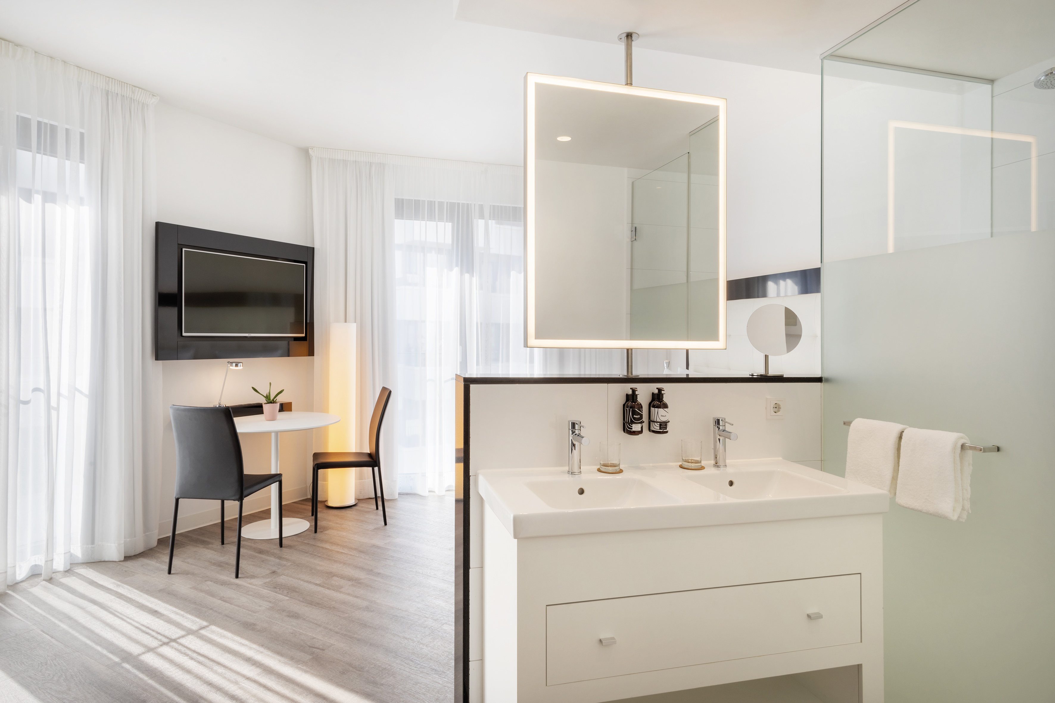 Modern hotel suite with dual vanity, lighted mirror, TV, and seating area.