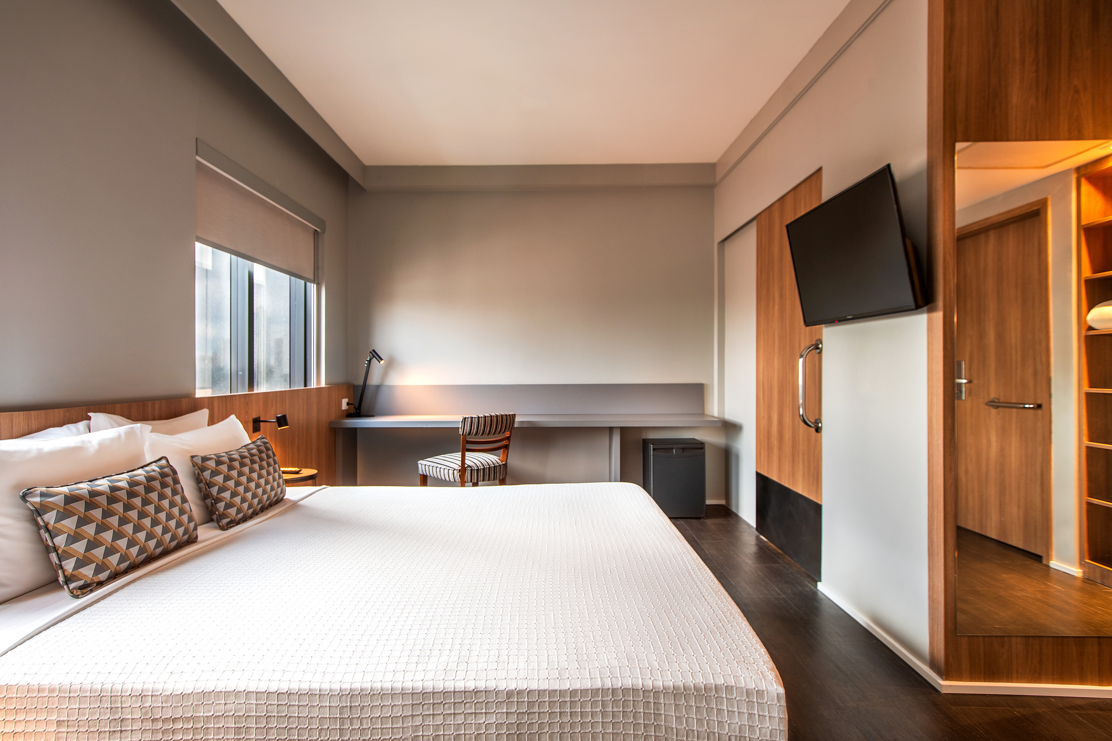 Modern hotel bedroom with white bed, patterned pillows, desk, and TV.