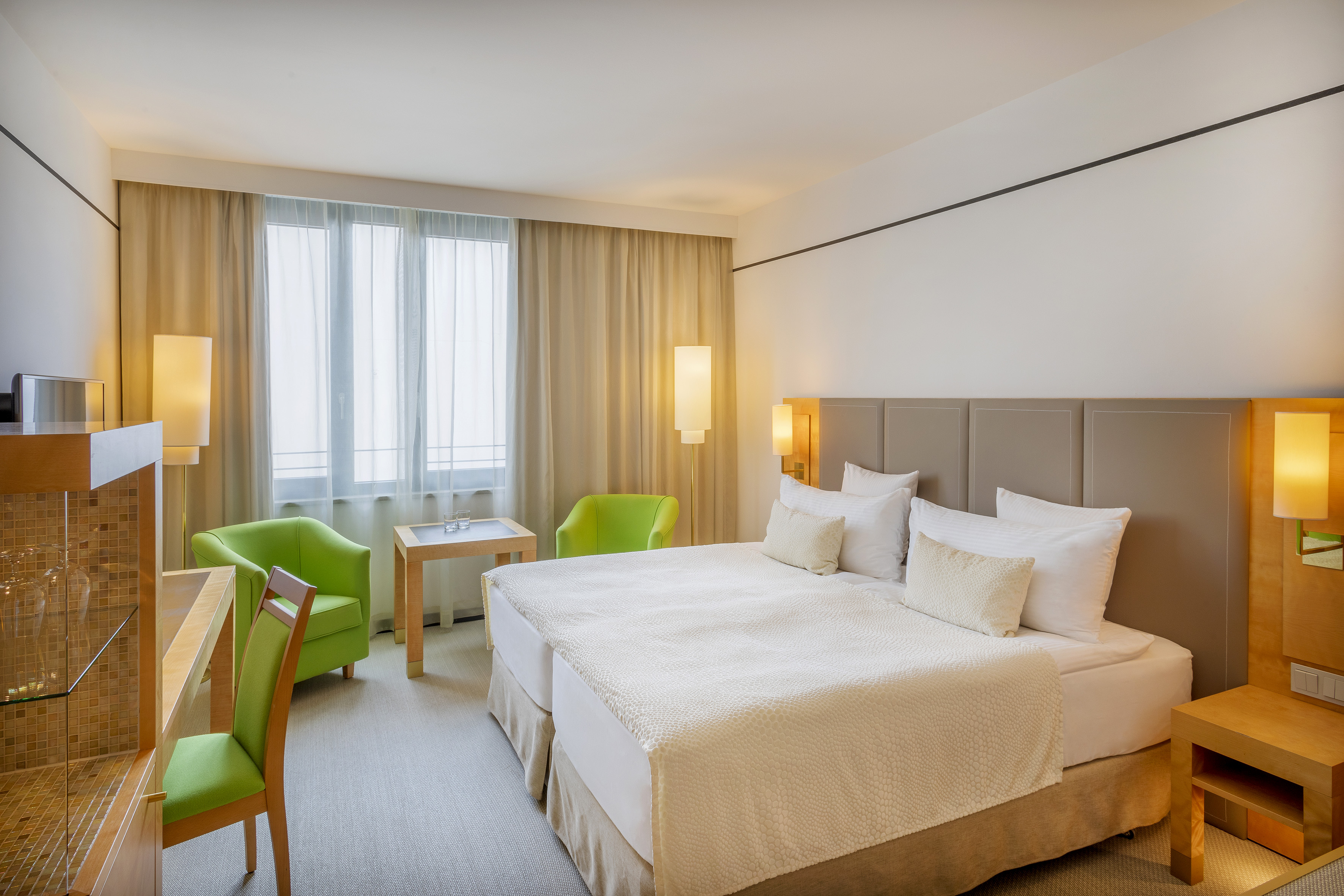 Modern hotel room with two beds, green armchairs, and bright windows.
