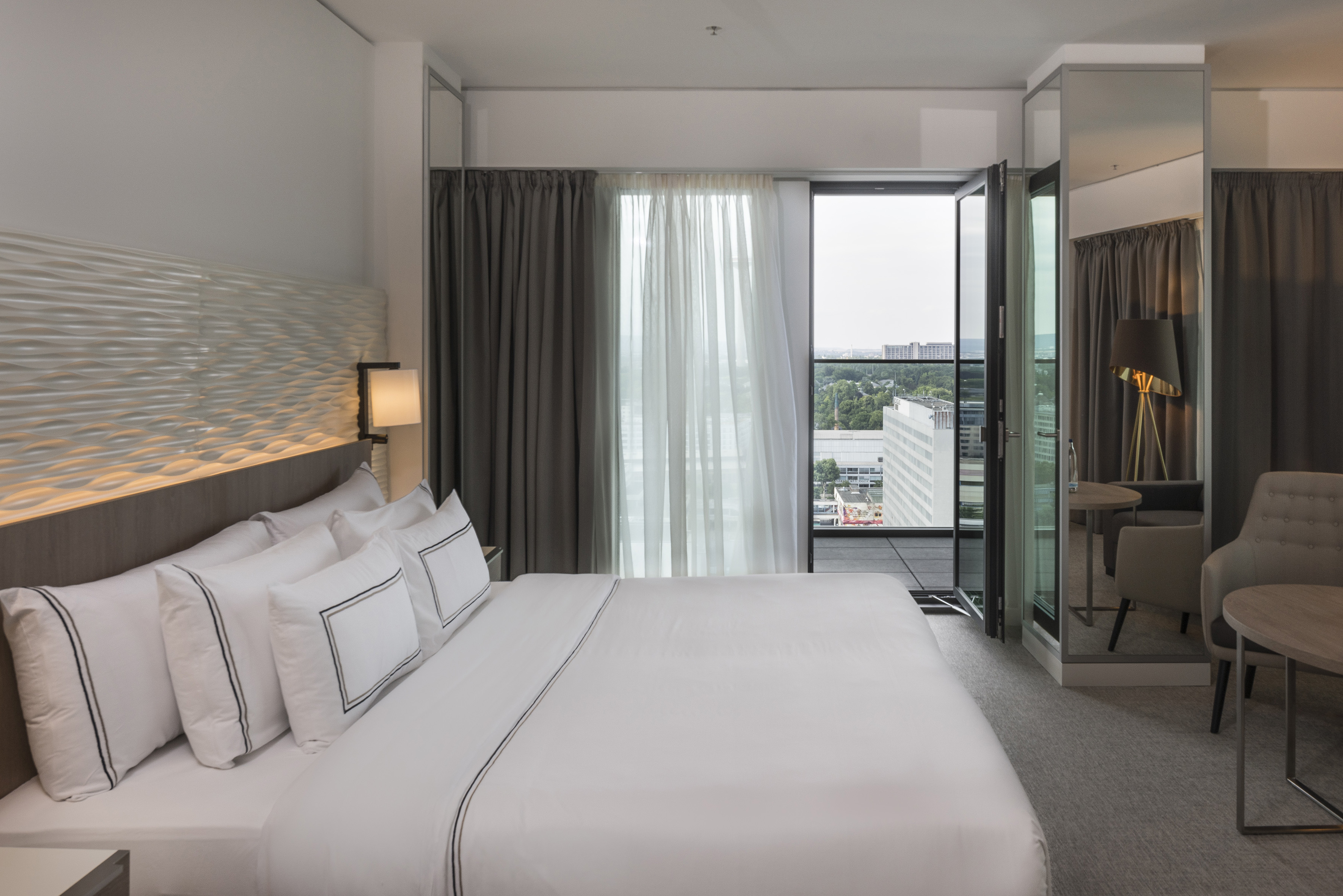 Spacious hotel room with white bed, city view balcony, and modern design.