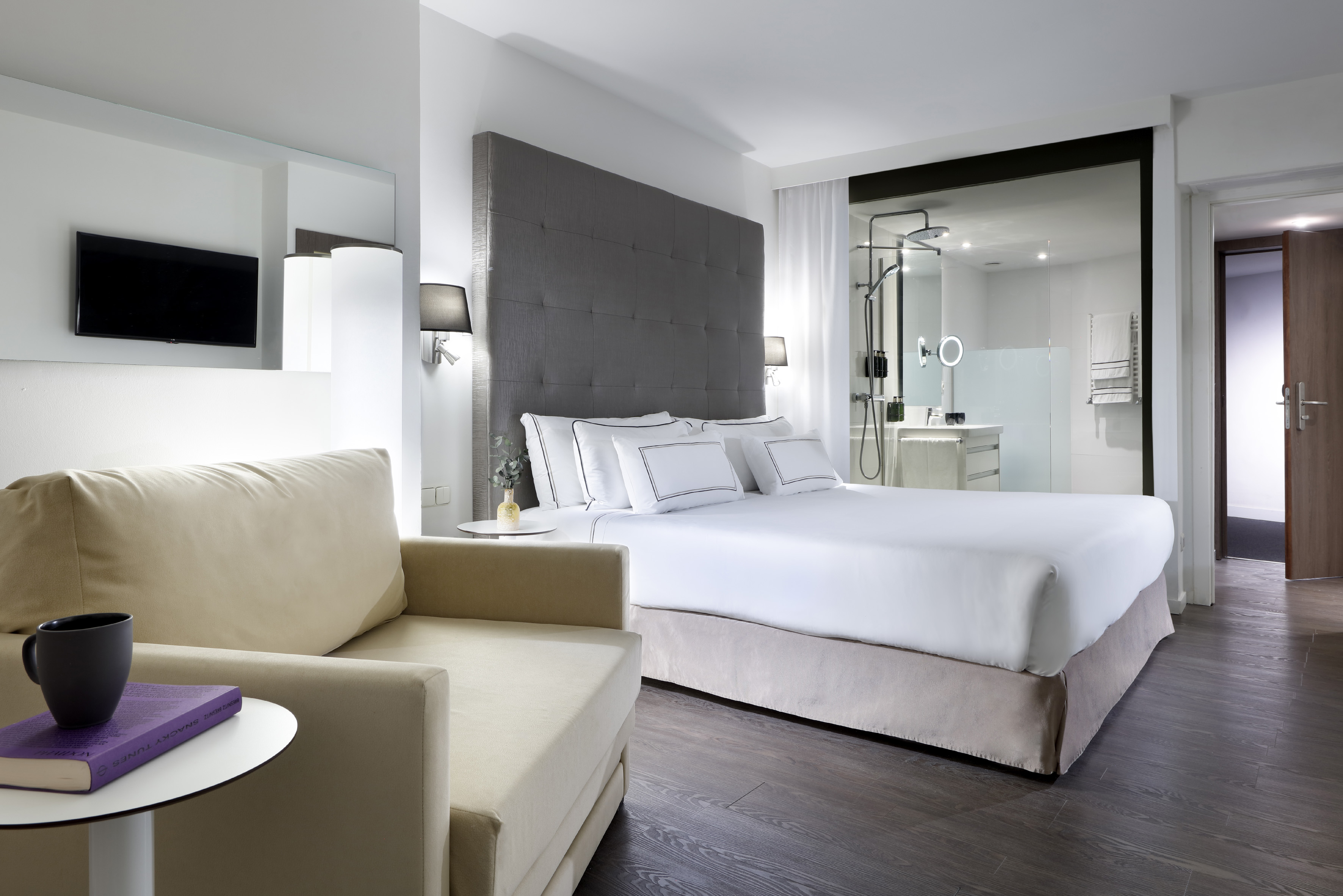 Sleek hotel bedroom with a king bed, chaise lounge, and glass-enclosed bathroom.