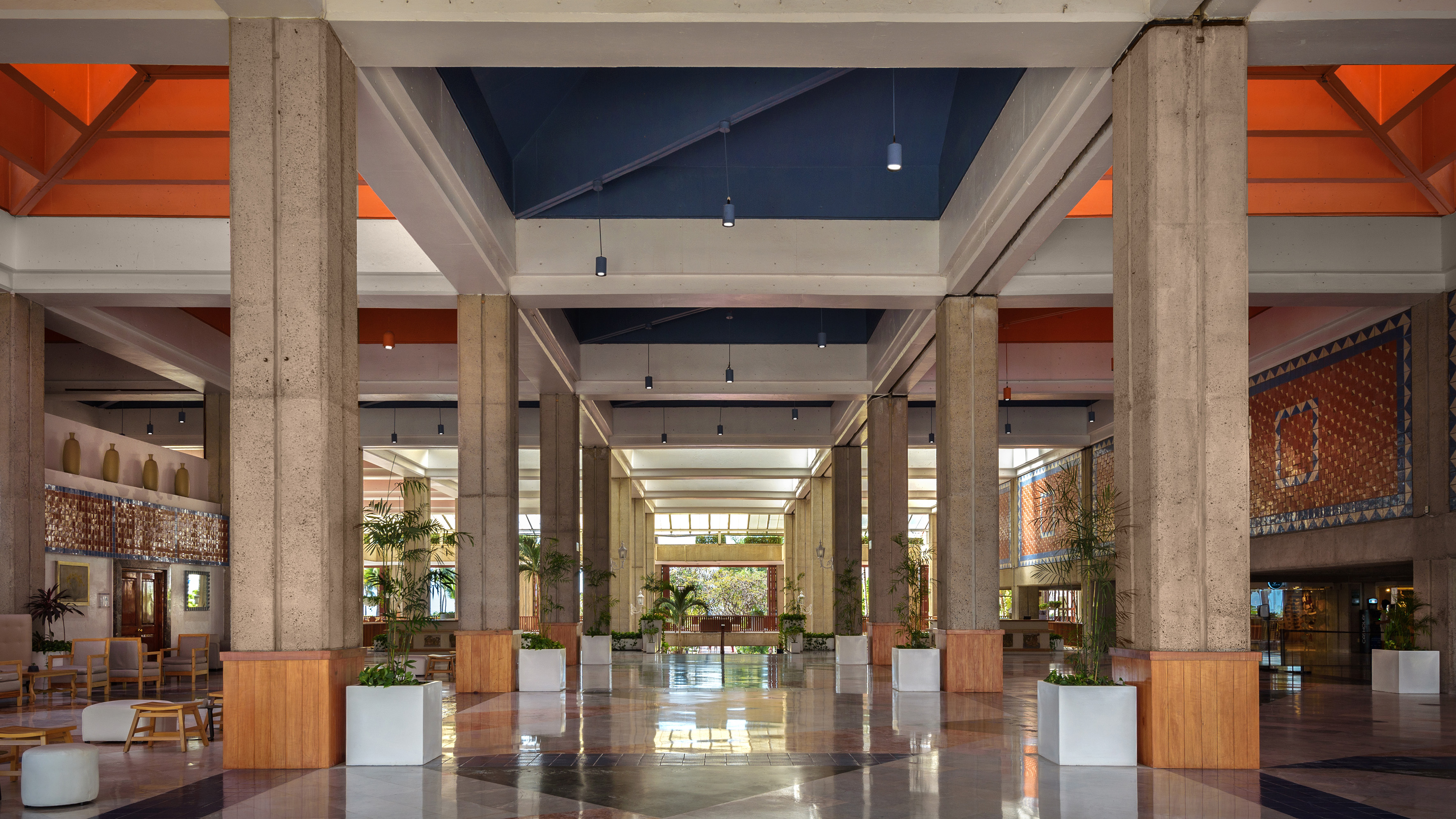 Spacious modern hotel lobby with concrete pillars, blue and orange ceilings, and patterned floor.