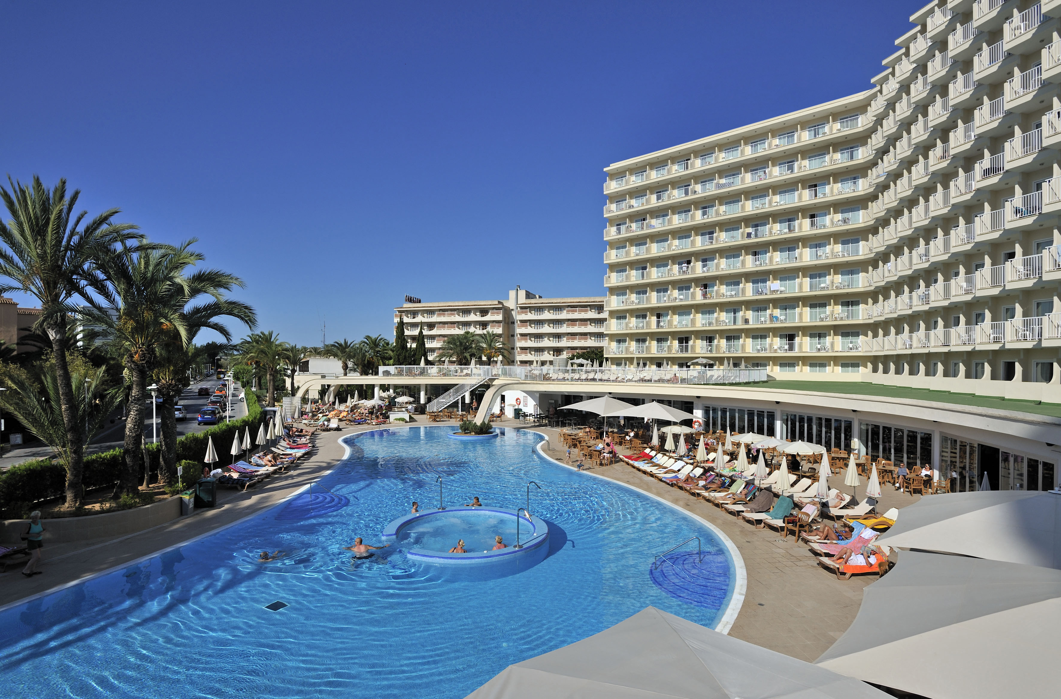 Sunny hotel resort with large swimming pool, sun loungers, and palm trees.