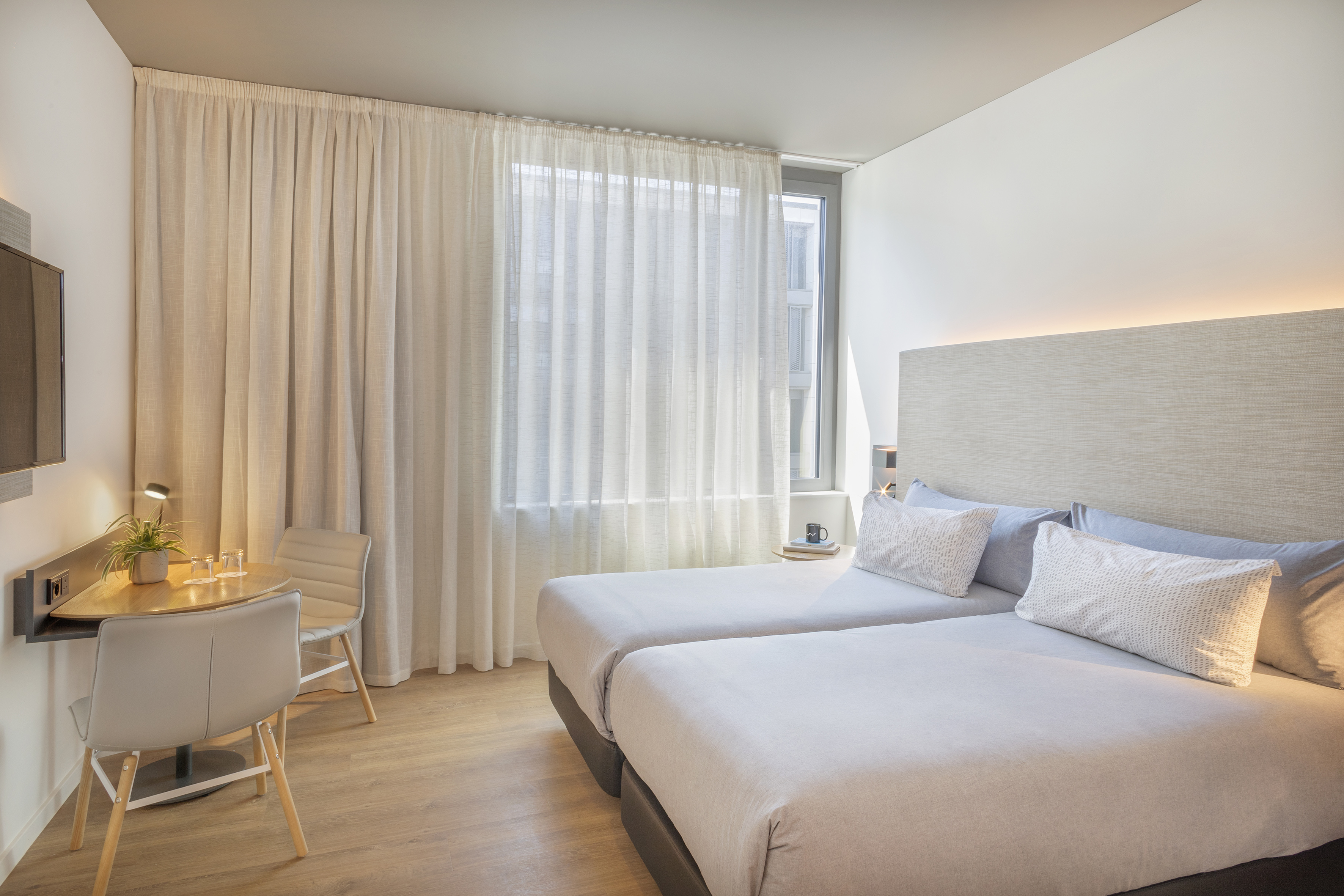 Modern hotel room with twin beds, desk, and sheer curtains.