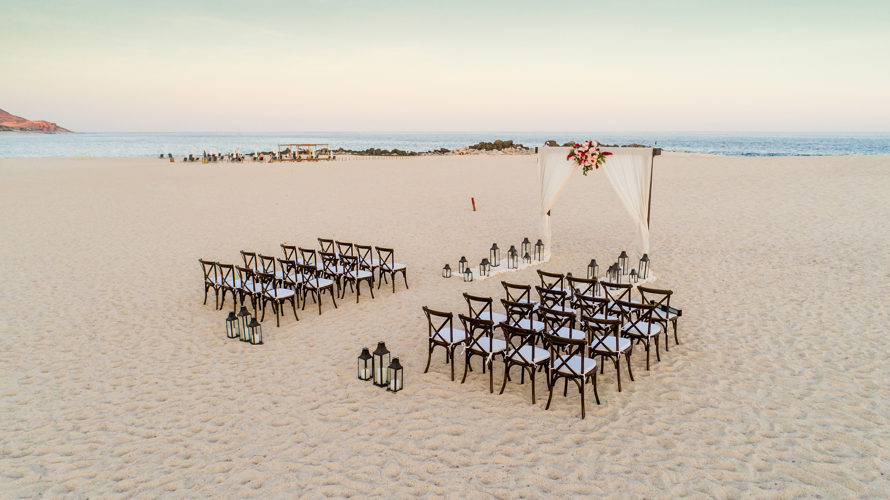Destination beach wedding setup with floral arch, chairs, and lanterns.