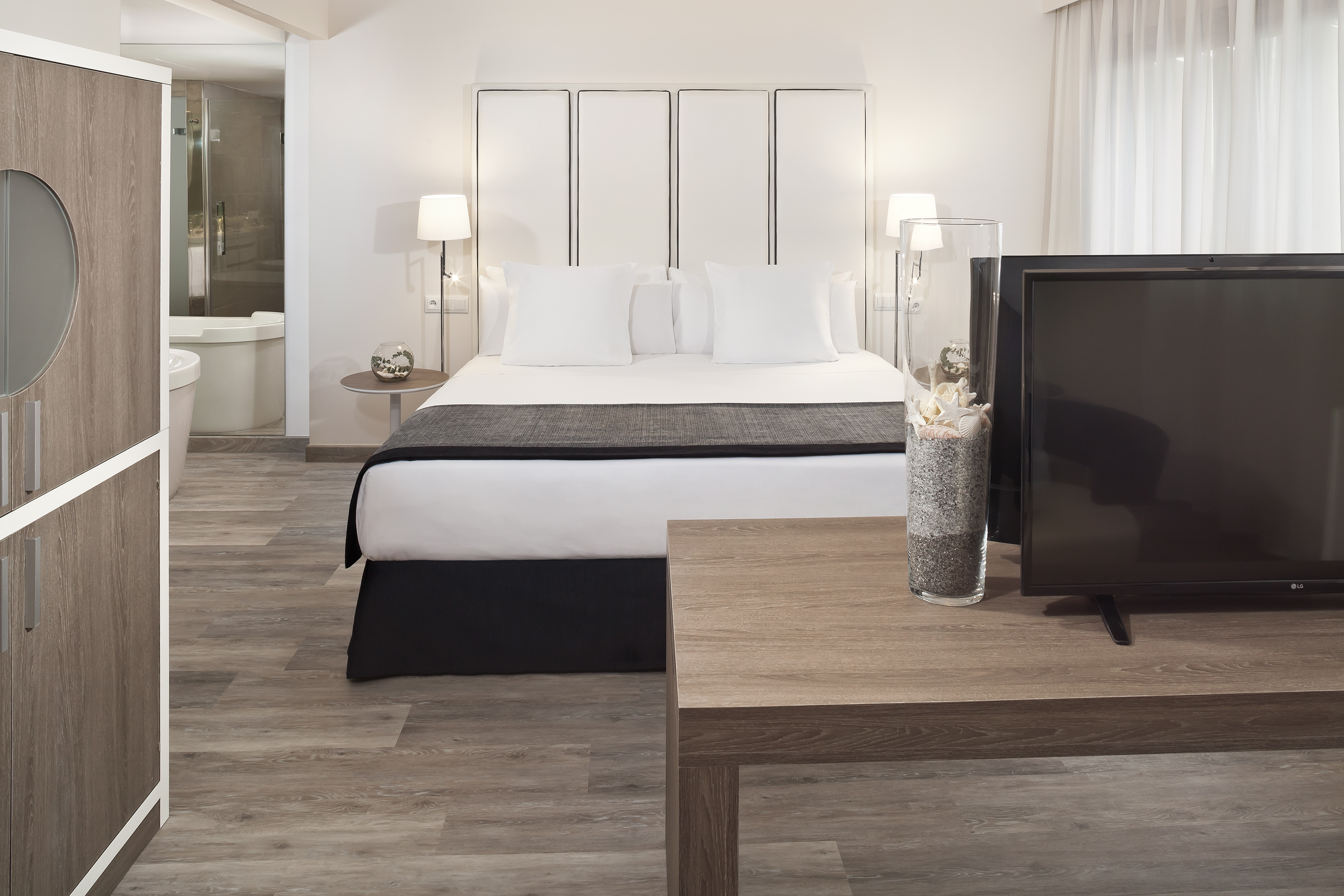 Modern hotel room with white bed, TV, wood flooring, and en-suite bathroom.