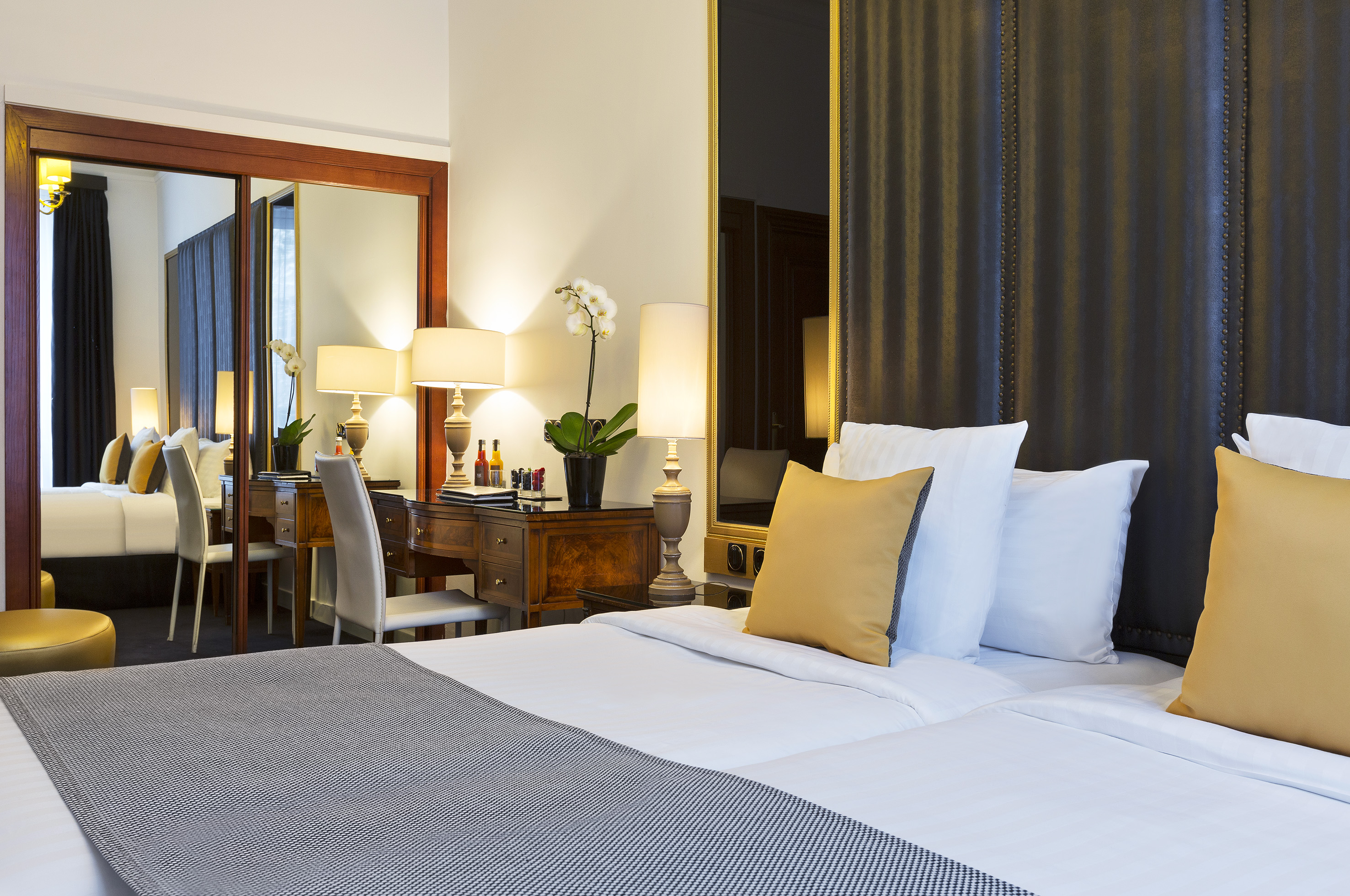 Luxury hotel room with large bed, golden pillows, desk with lamps and orchid, mirror.