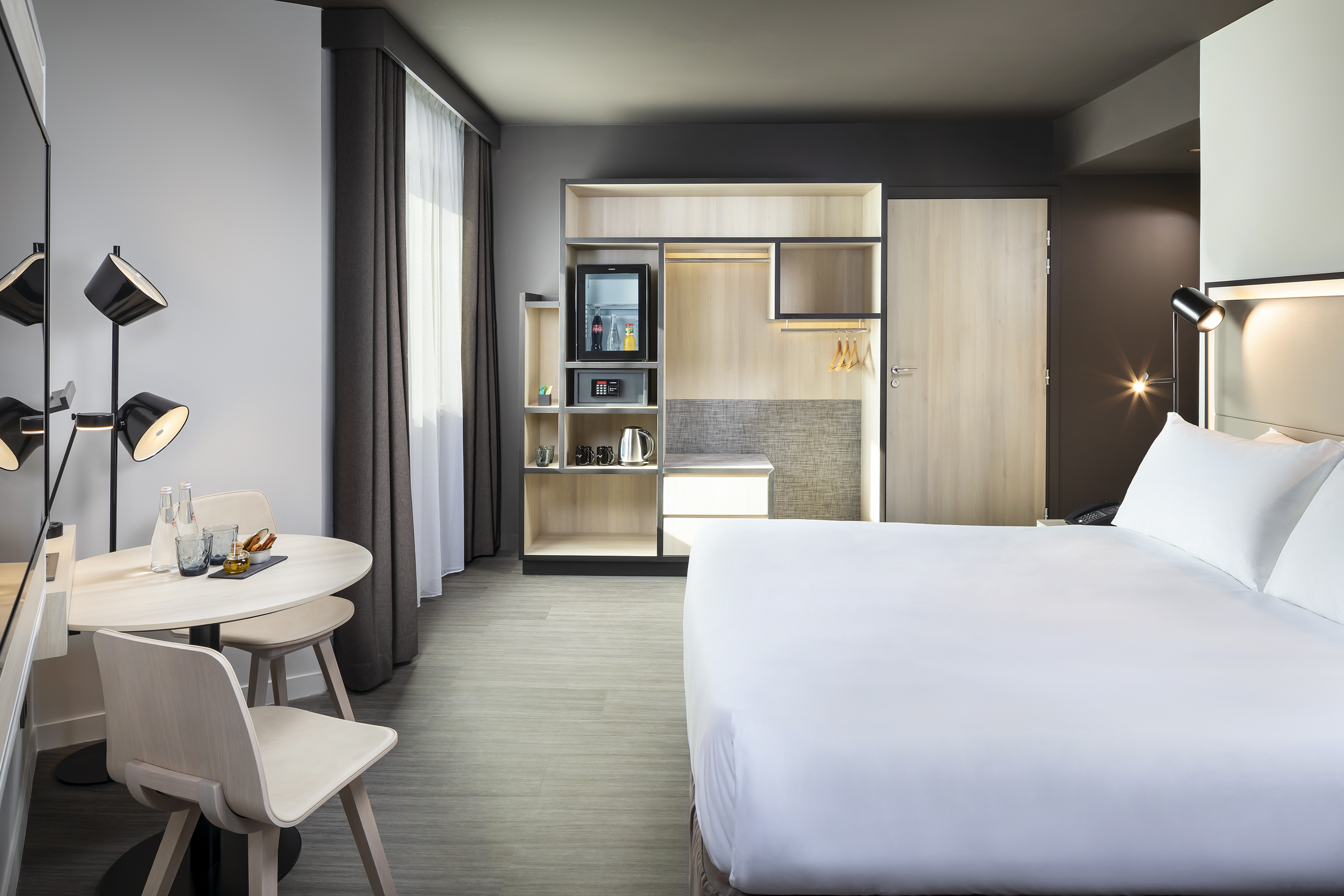 Modern hotel room with bed, dining table, mini-fridge, and wardrobe.