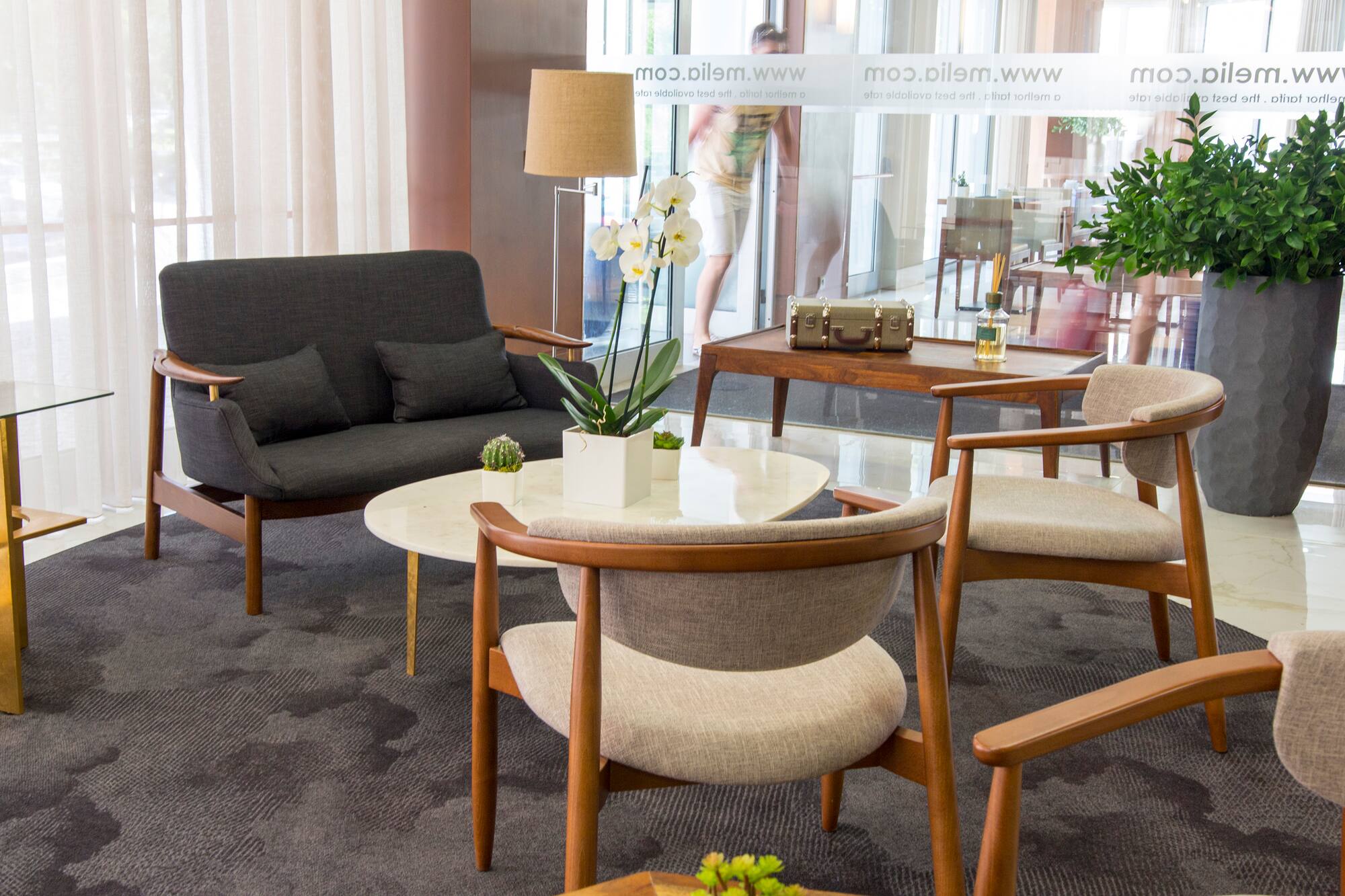 Modern hotel lobby with grey sofa, wood chairs, white orchids, and patterned rug.
