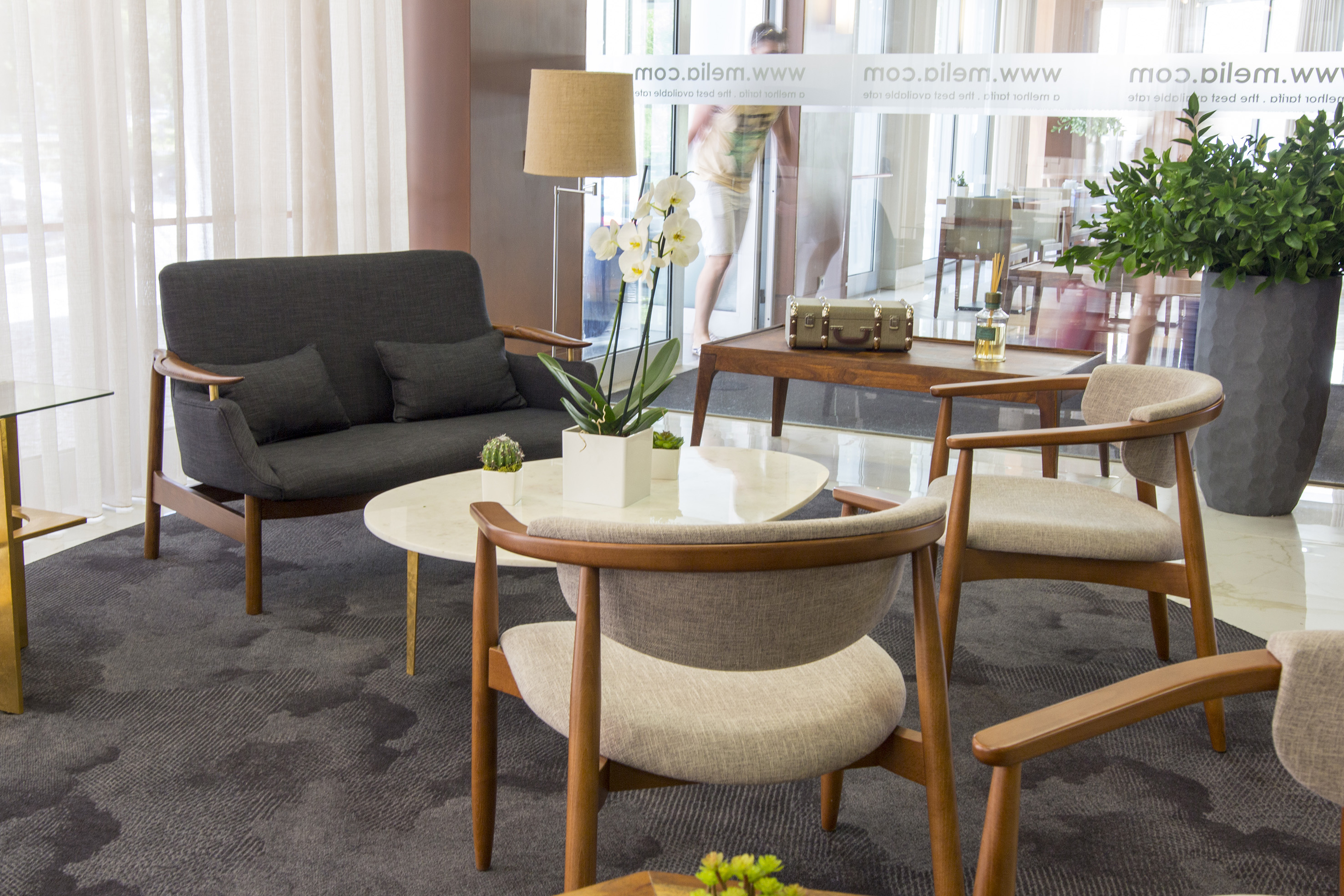 Modern hotel lobby with grey sofa, wood chairs, white orchids, and patterned rug.