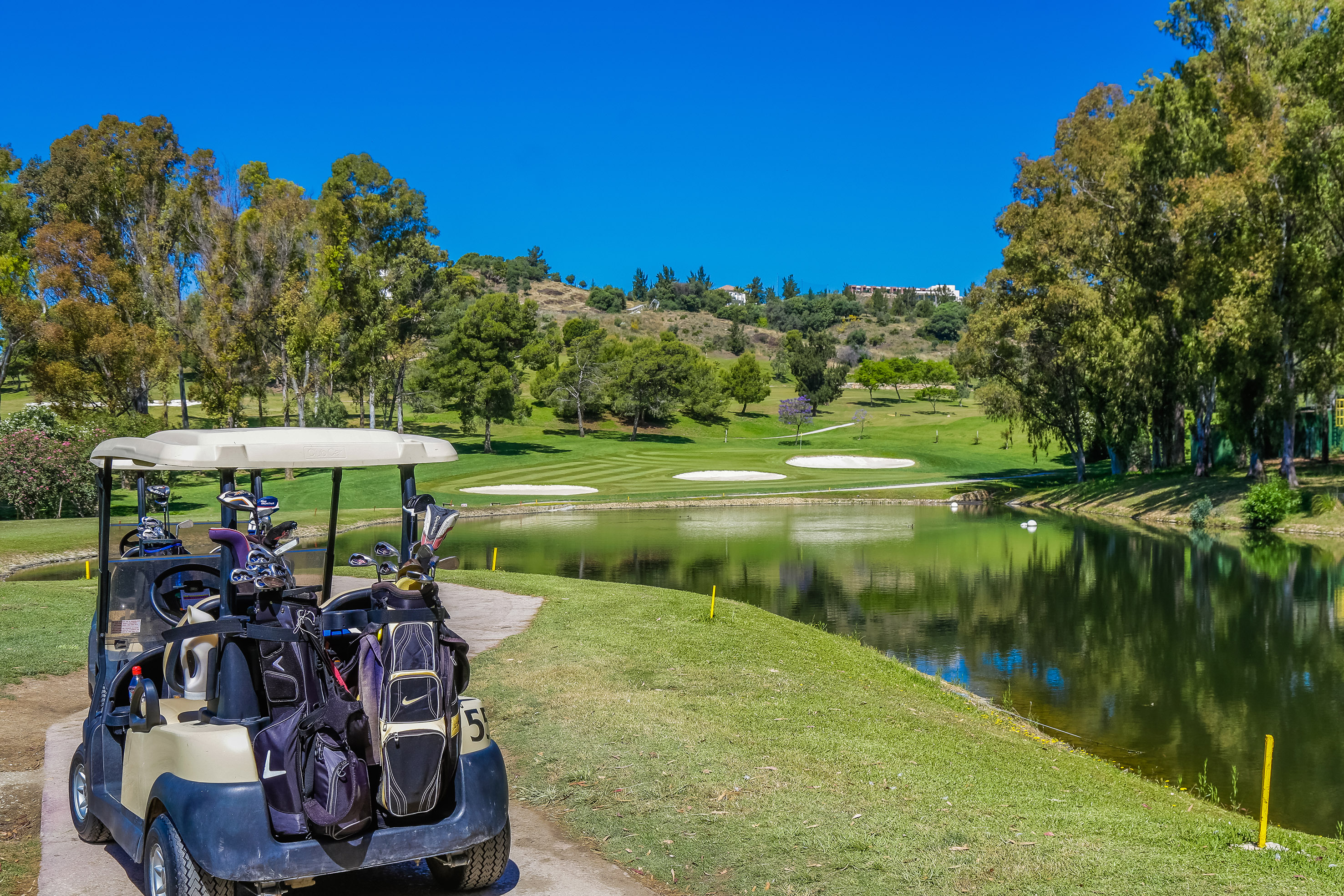 Golf cart with clubs by a lake on a sunny golf course with bunkers.