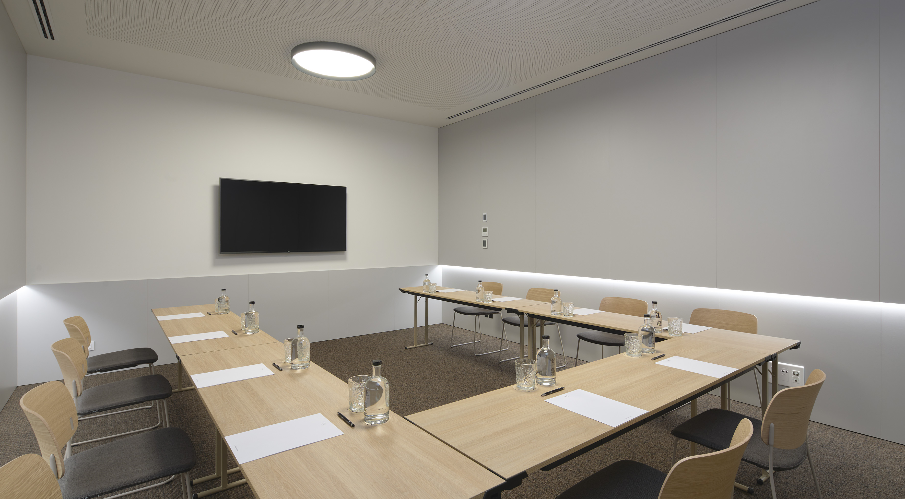 Modern conference room with U-shaped tables, grey chairs, and wall-mounted screen.
