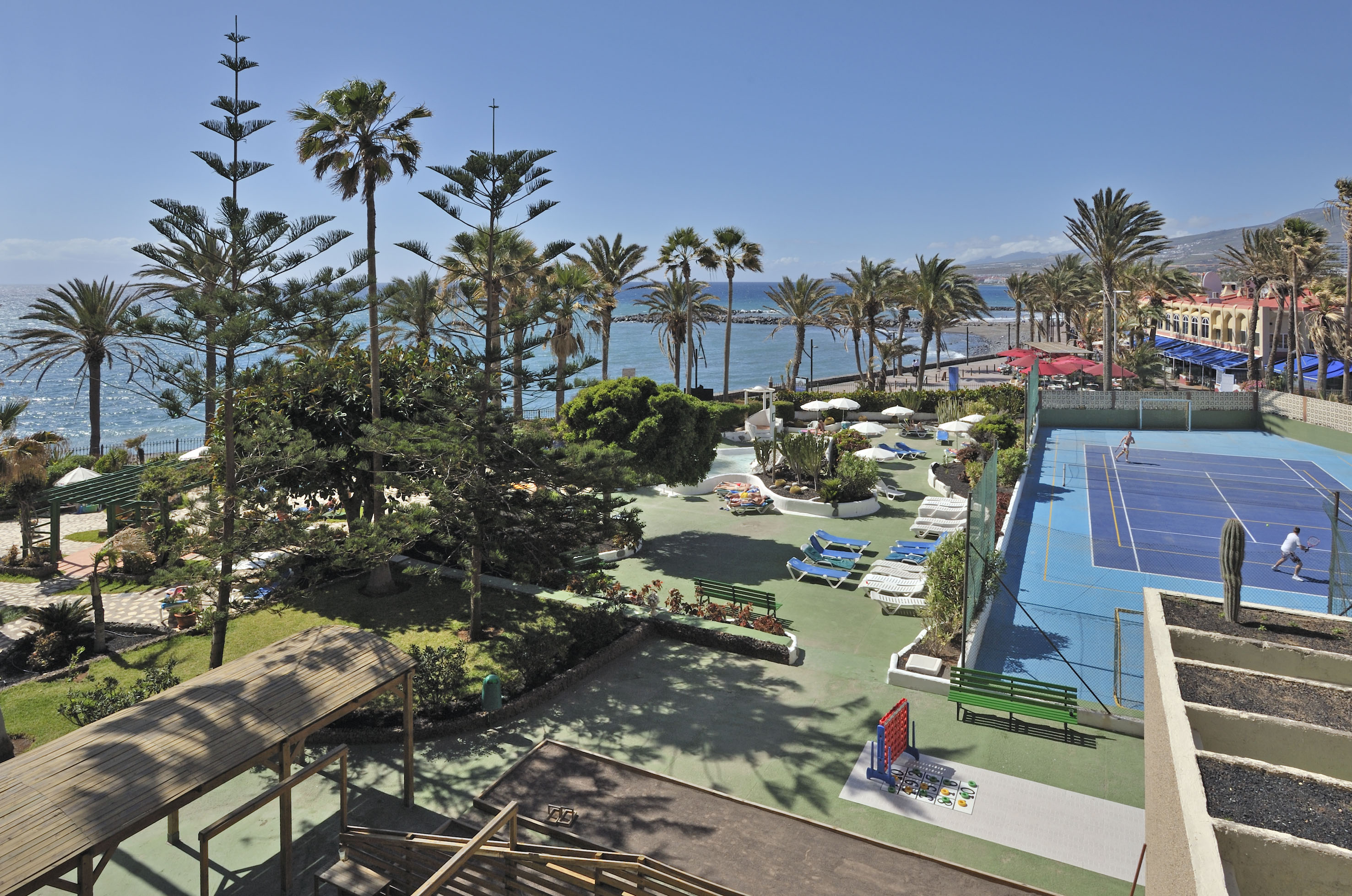 Oceanfront resort with tennis court, swimming pool, and palm trees under blue sky.