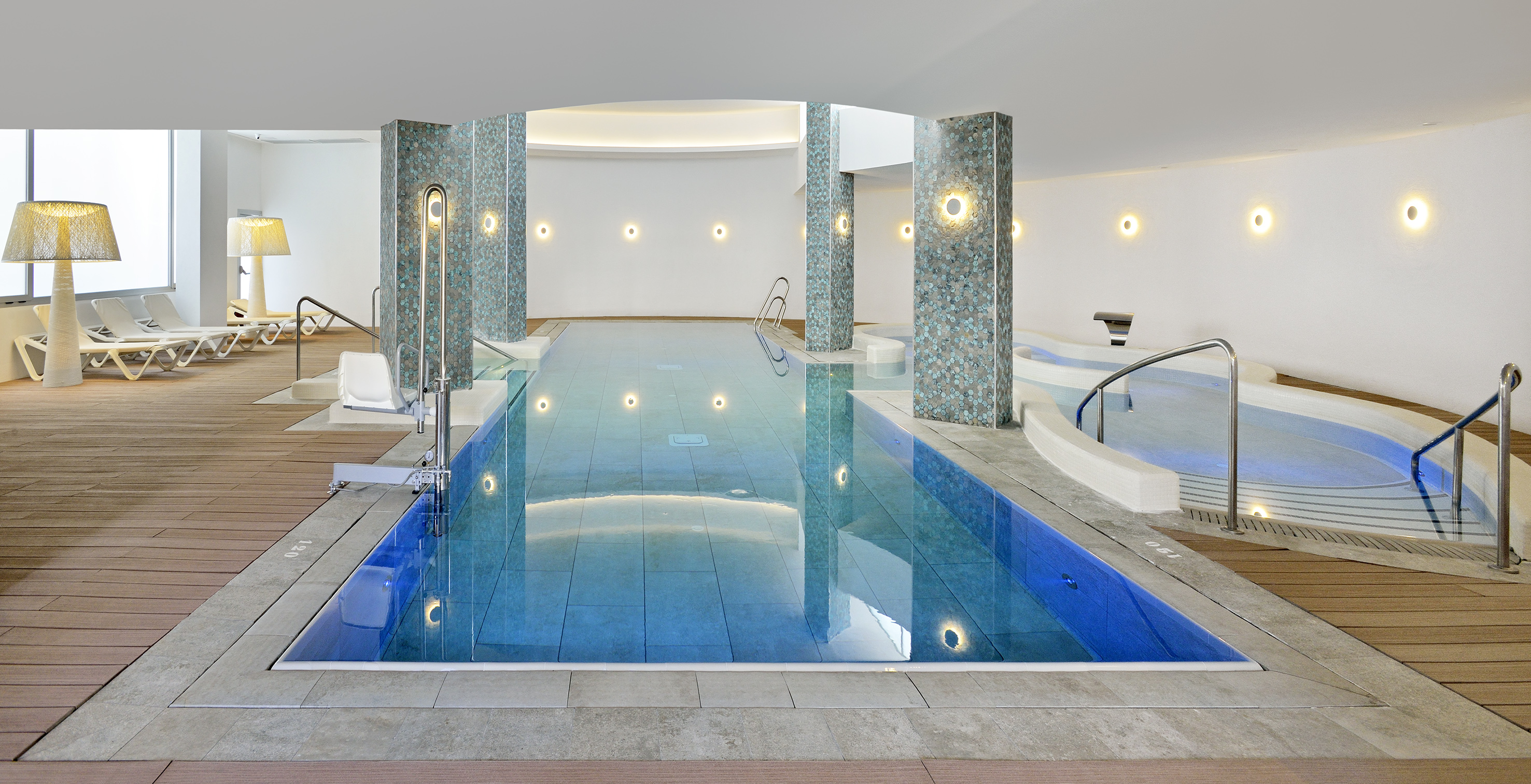 Modern indoor swimming pool with hot tub, loungers, and accessibility lift.