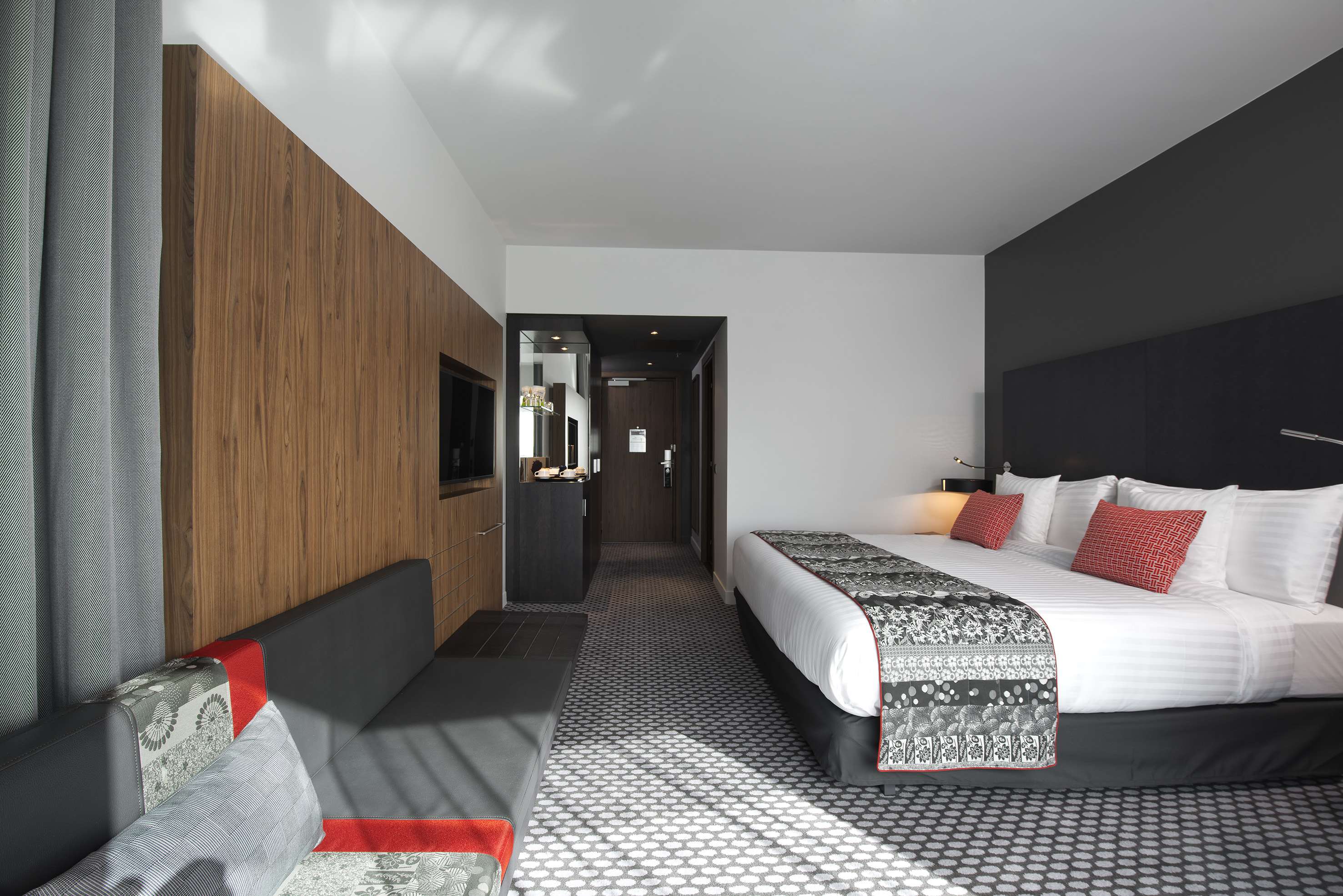 Modern hotel room with king bed, sofa, wood paneling, and patterned carpet.