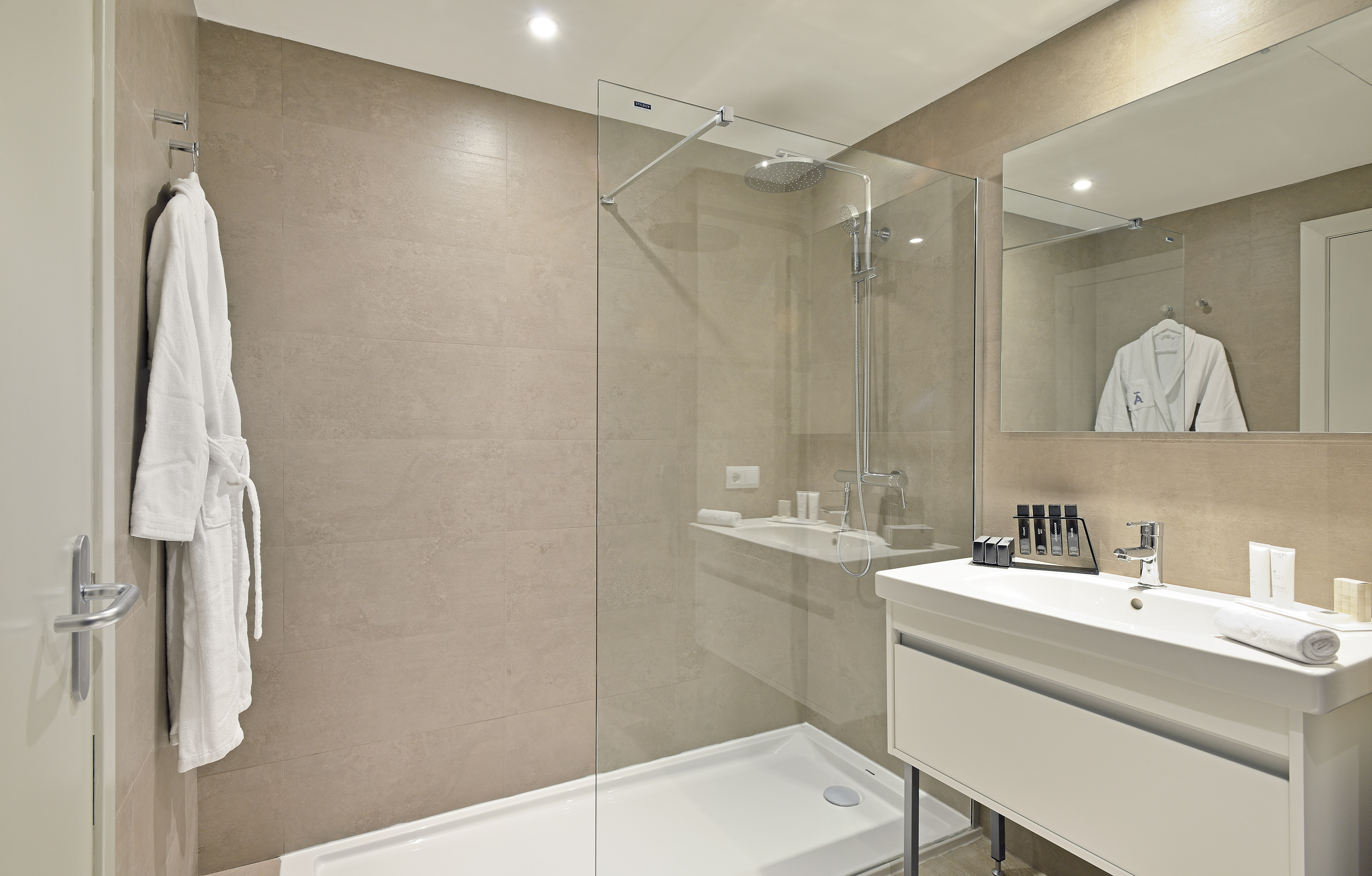 Modern hotel bathroom with glass shower, white vanity, and beige tiled walls.