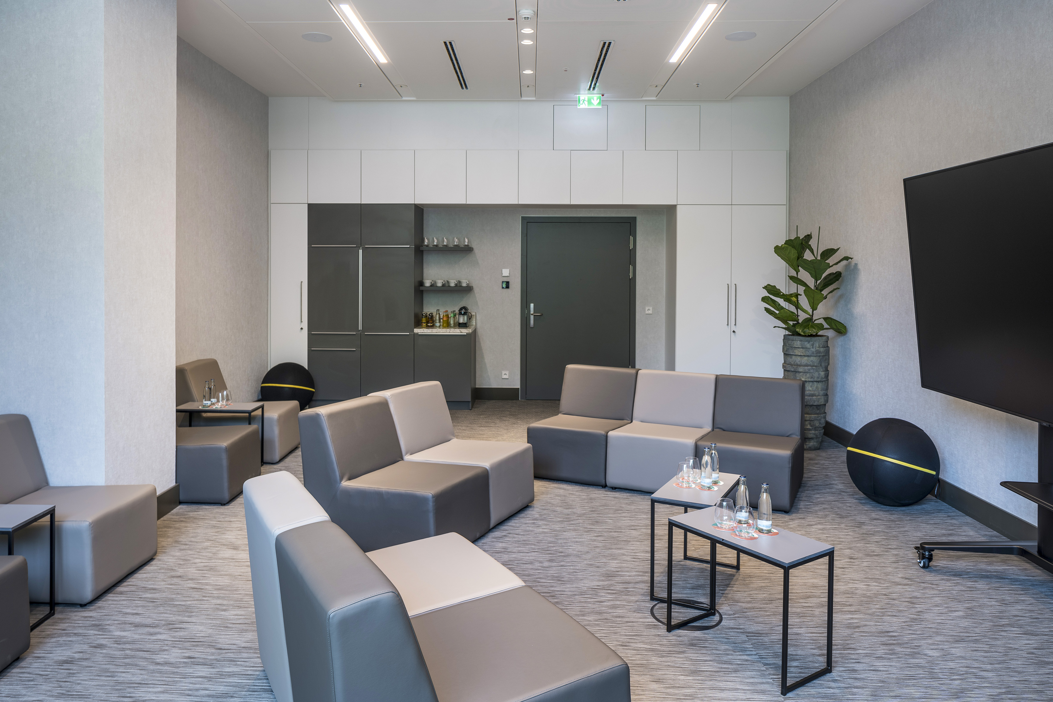 Modern corporate lounge with modular grey seating, kitchenette, and plant.