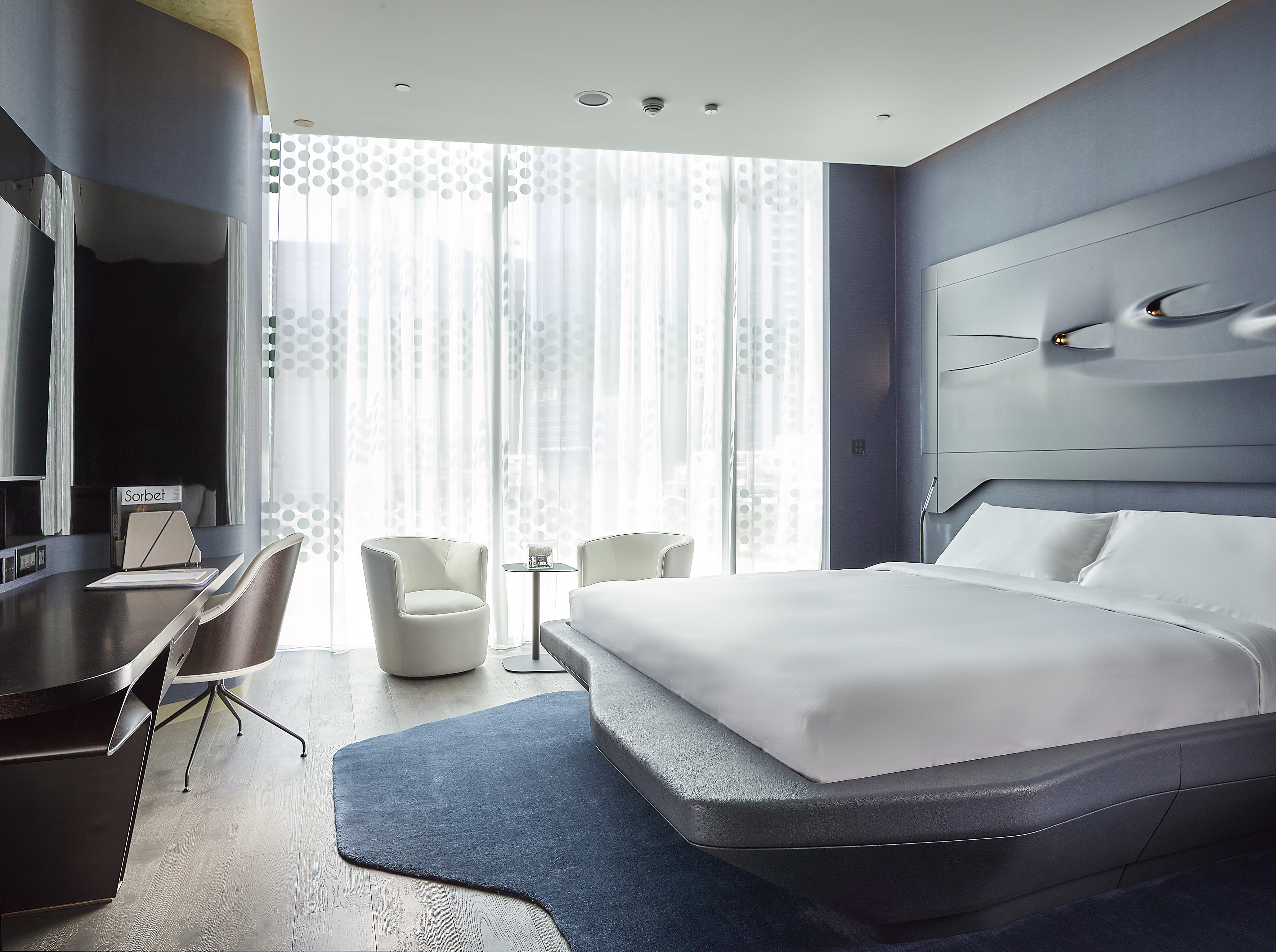 Luxury hotel bedroom with modern bed, blue headboard, desk, and window.