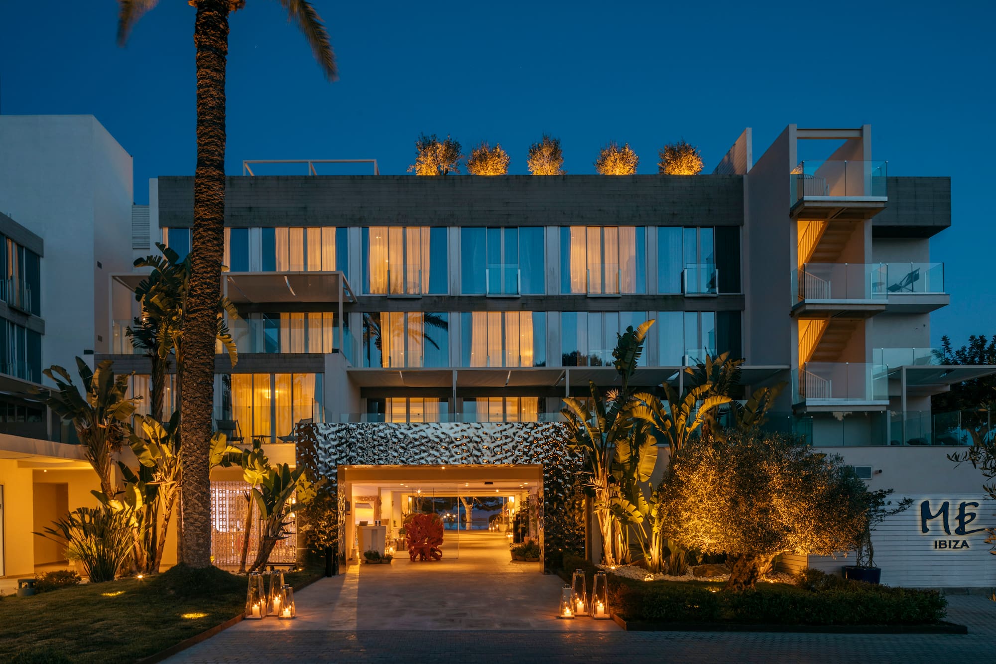 Modern ME Ibiza hotel entrance at dusk with lit palm trees and lanterns.