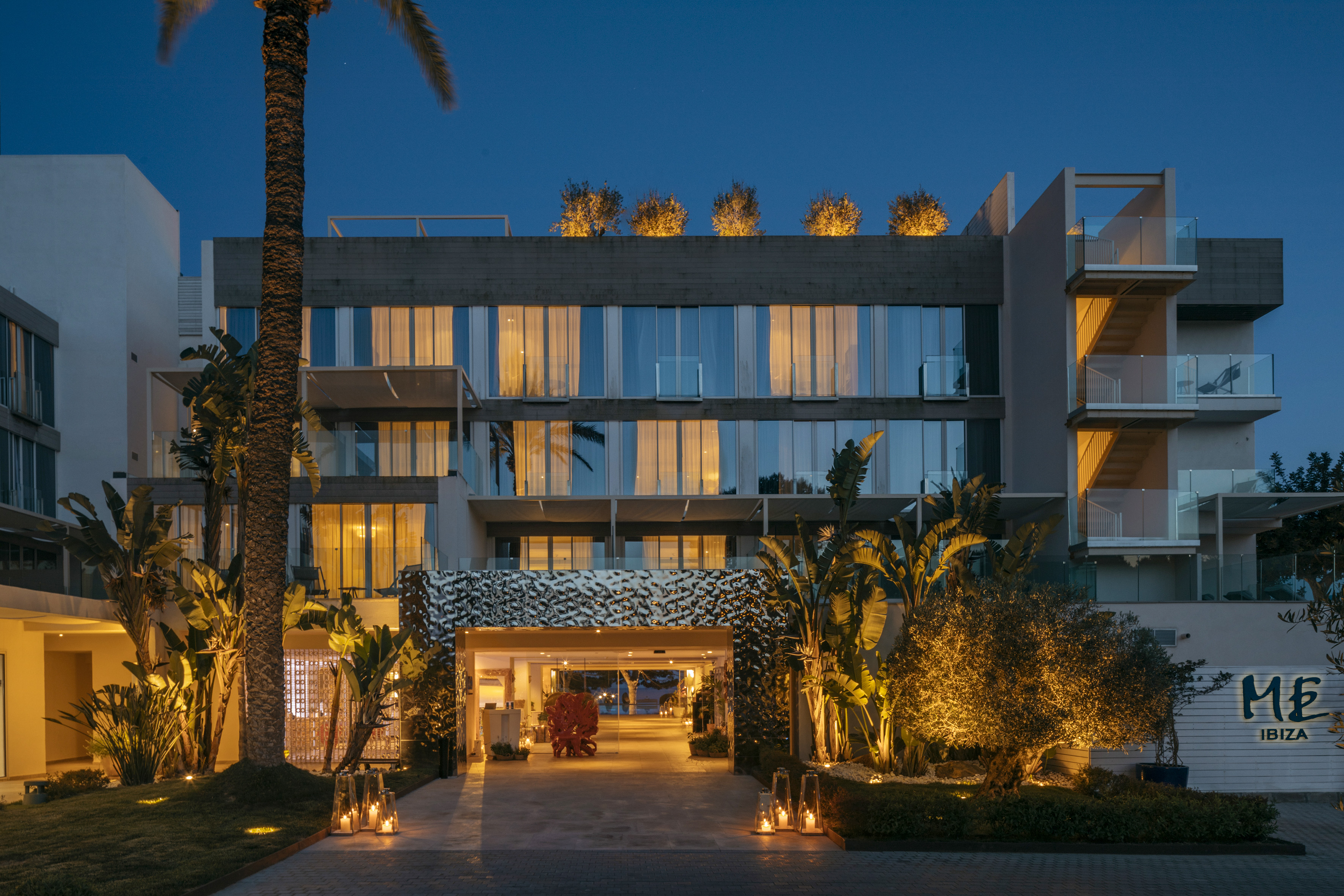 Modern ME Ibiza hotel entrance at dusk with lit palm trees and lanterns.