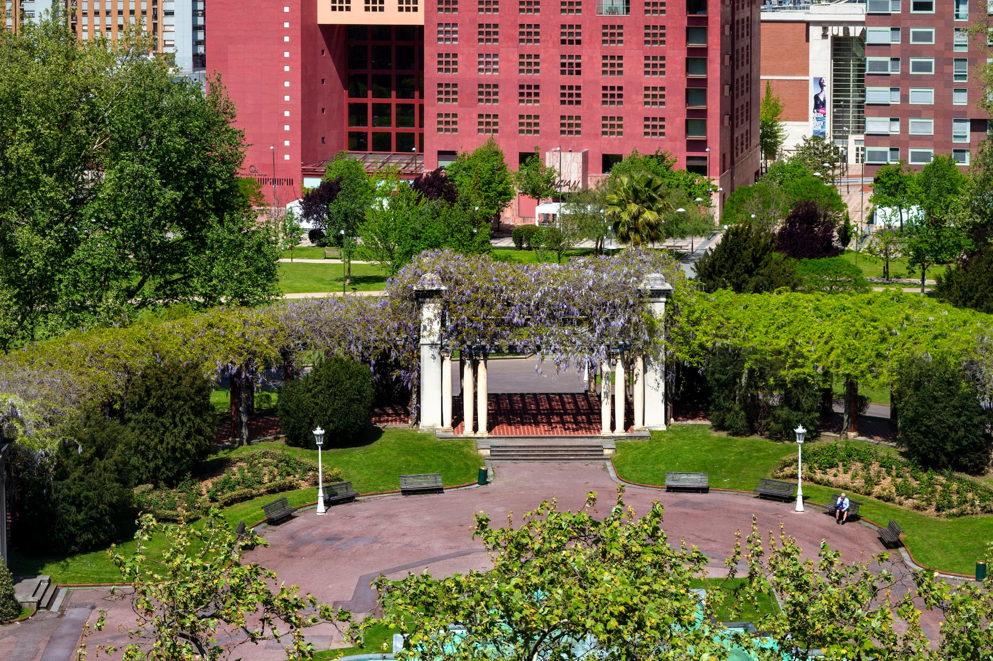 Bilbao park with wisteria pergola, fountain, and the red Melia hotel building.