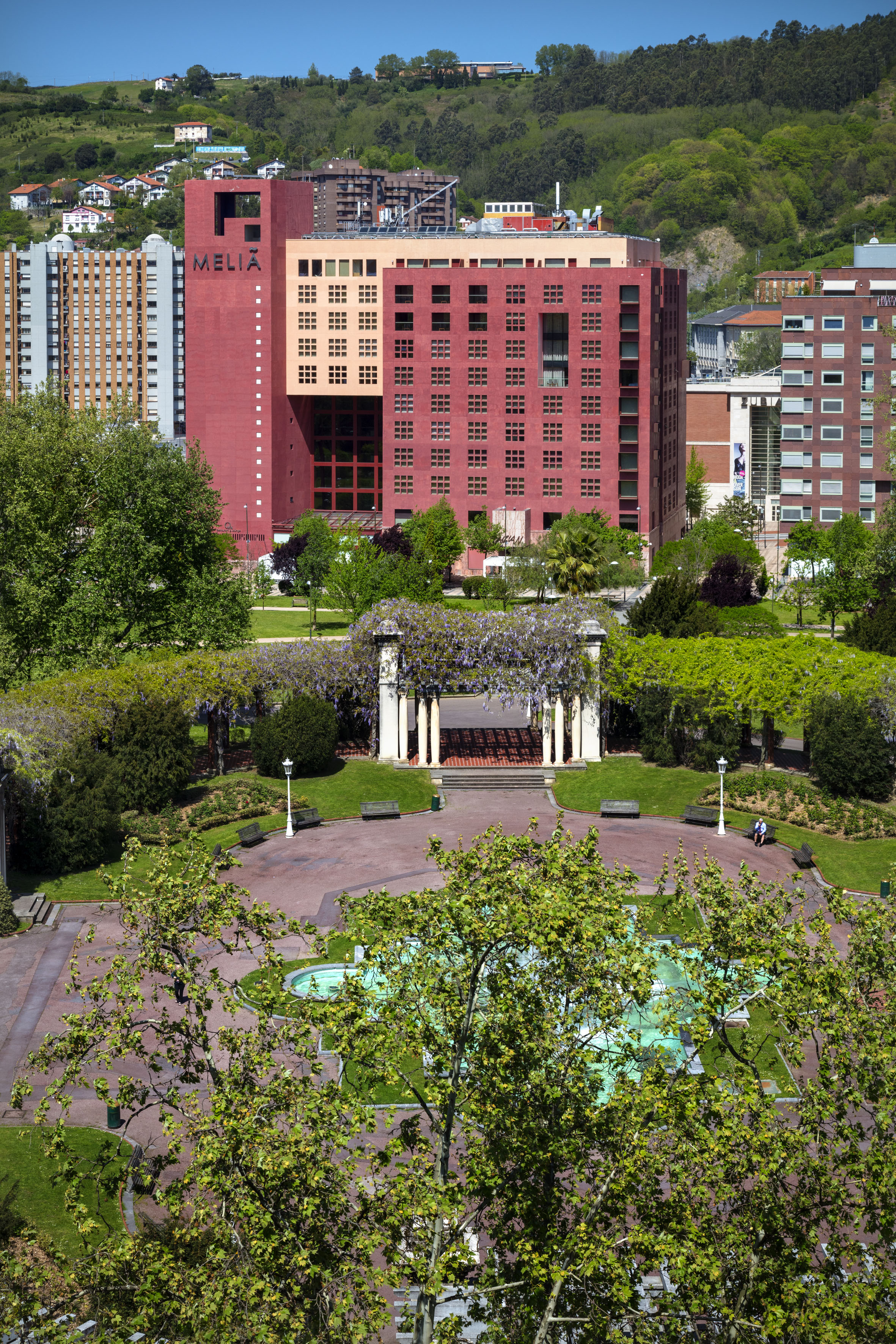 Bilbao park with wisteria pergola, fountain, and the red Melia hotel building.