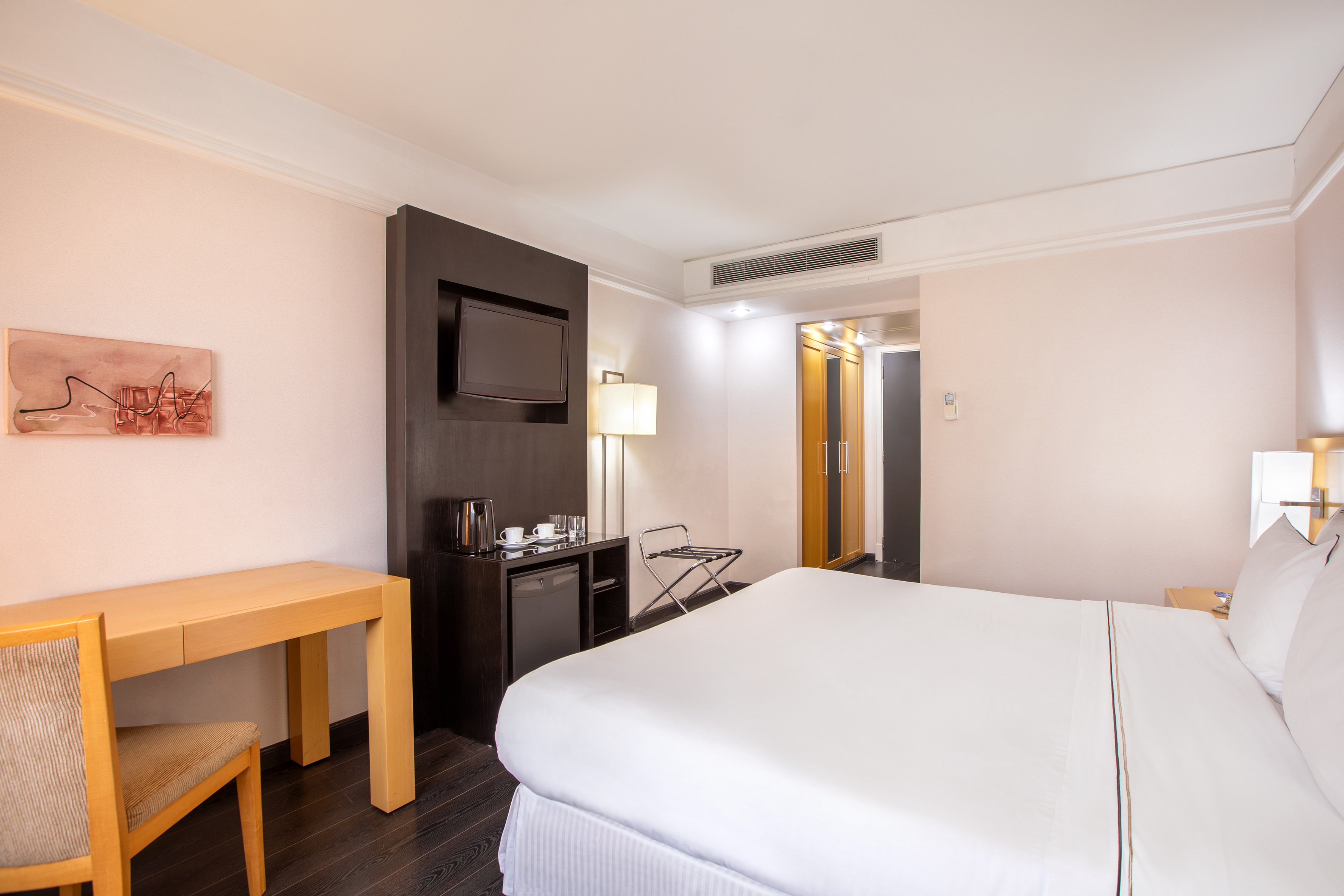 Modern hotel guest room with bed, TV, desk, and minibar.