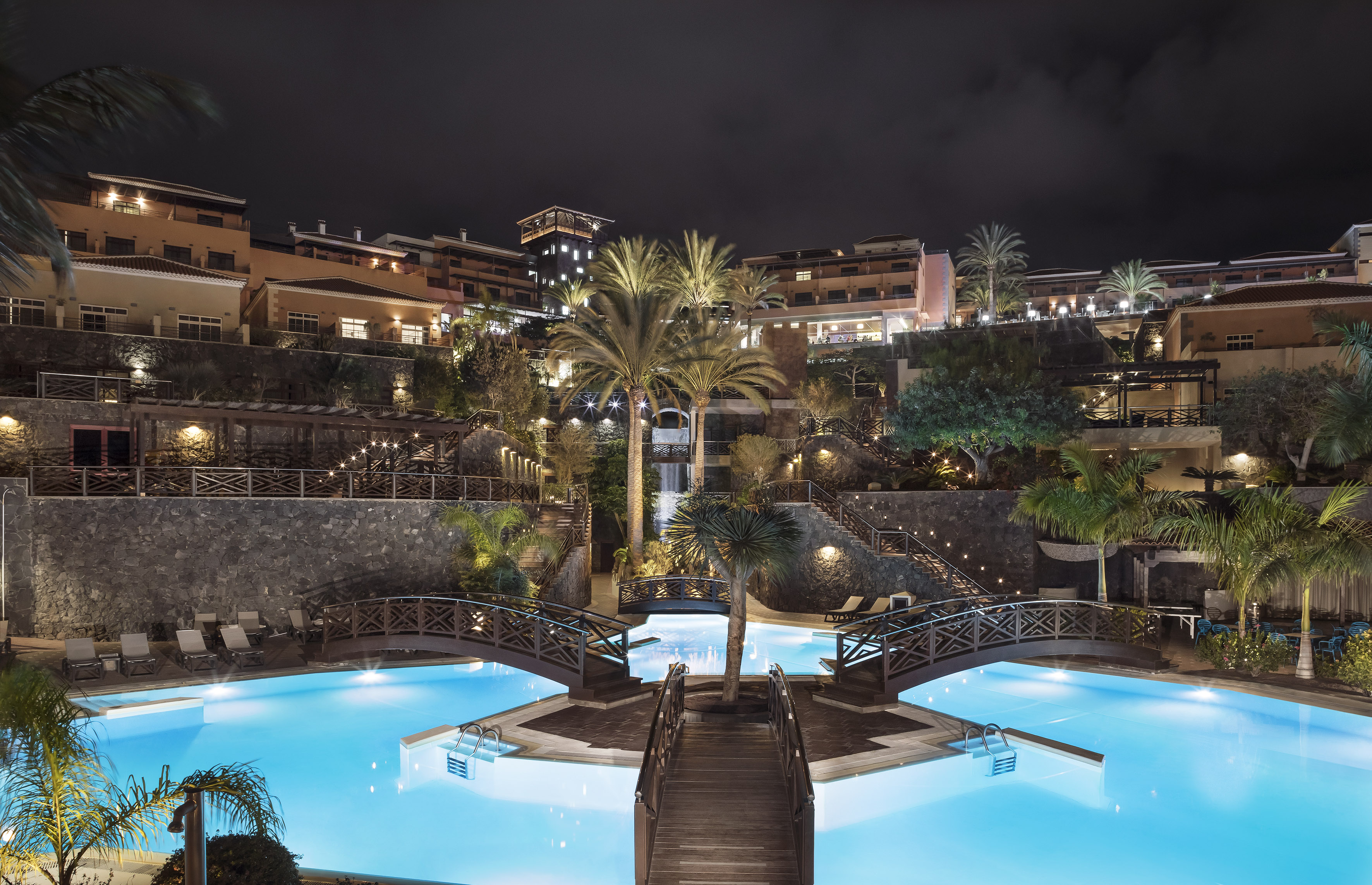 Luxury resort pool at night with illuminated palm trees, bridges, and buildings.