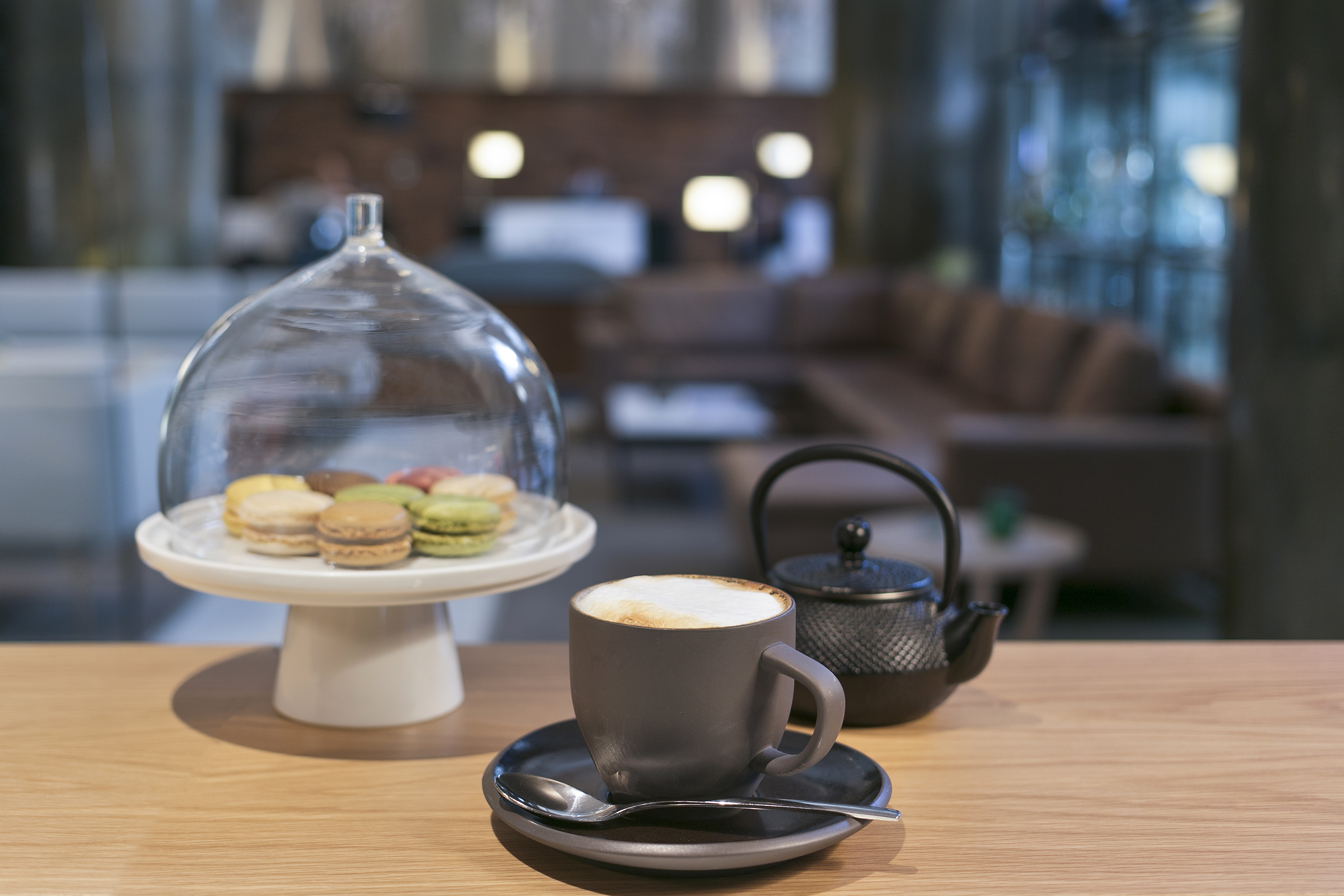 Colorful macarons under cloche, cappuccino, and teapot on wooden table.