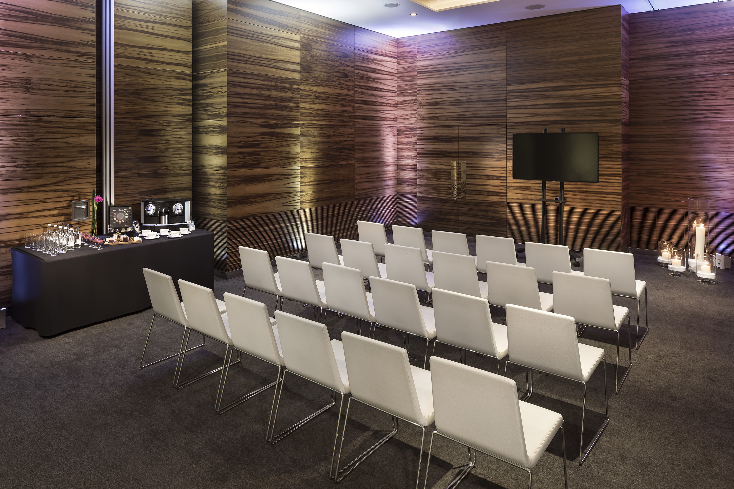 Modern conference room with white chairs, dark wood walls, and refreshment bar.