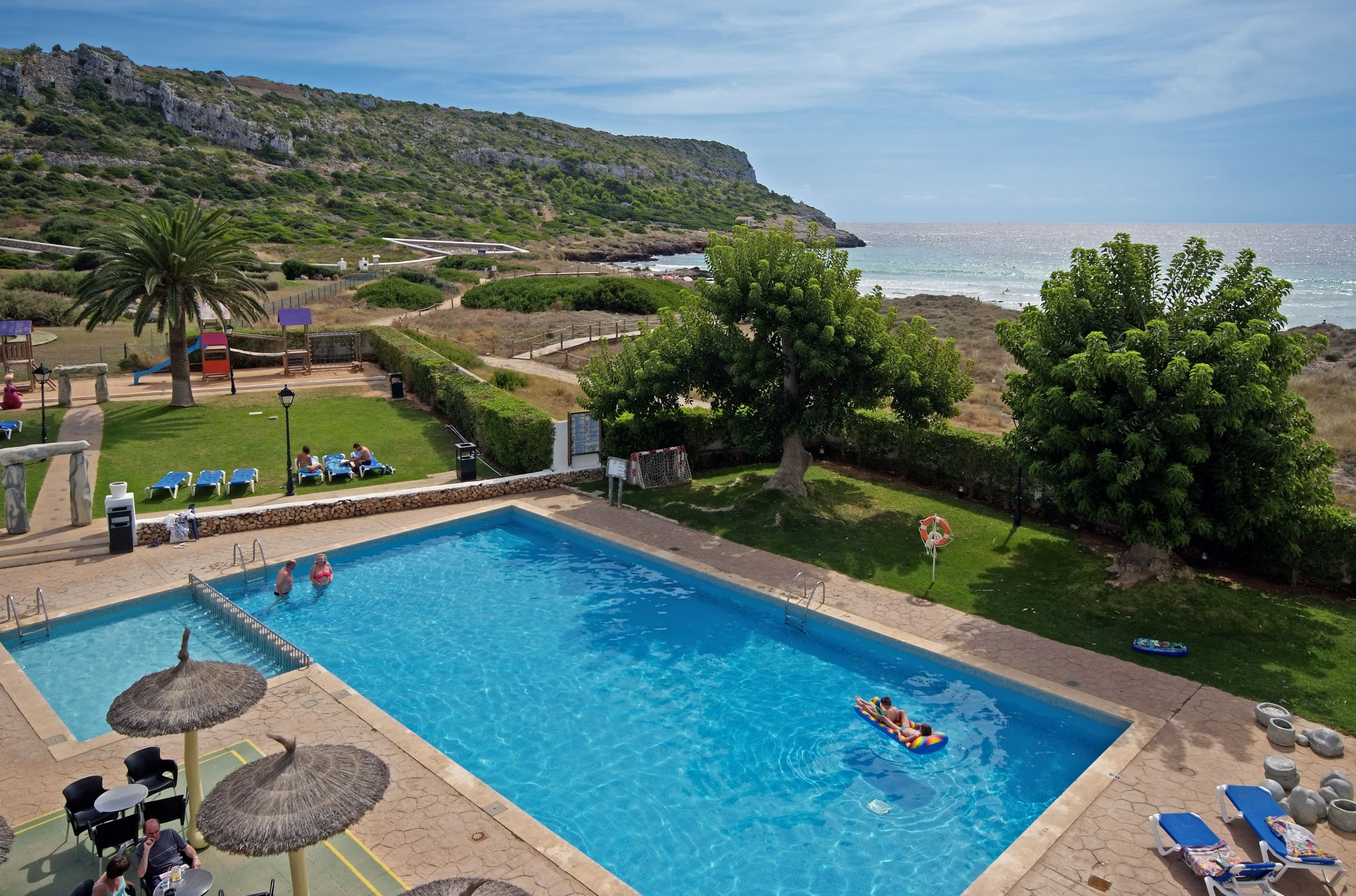 Resort swimming pool with guests, palm trees, green hills, and ocean view.