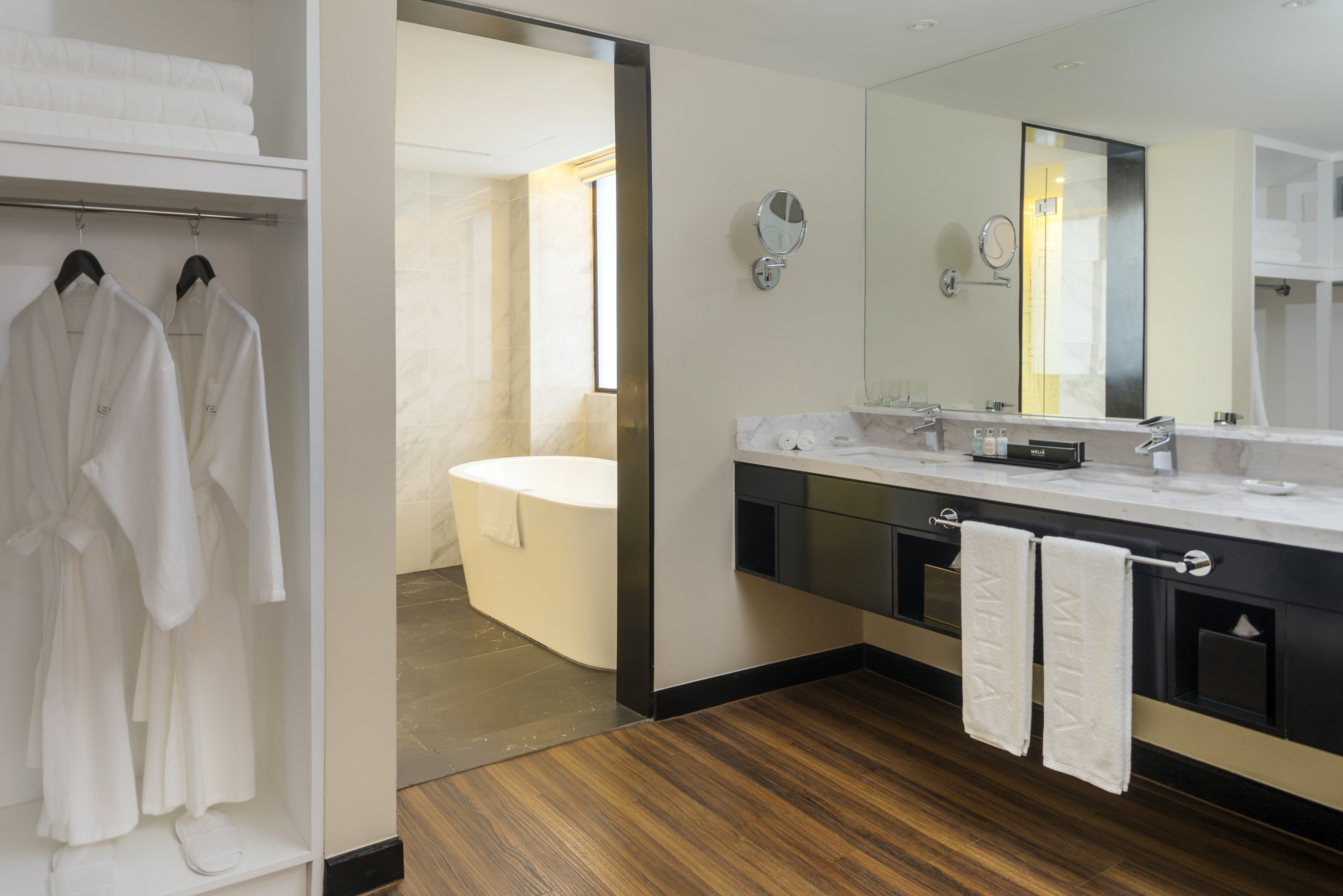 Modern hotel bathroom featuring double vanity, bathtub, and plush robes.