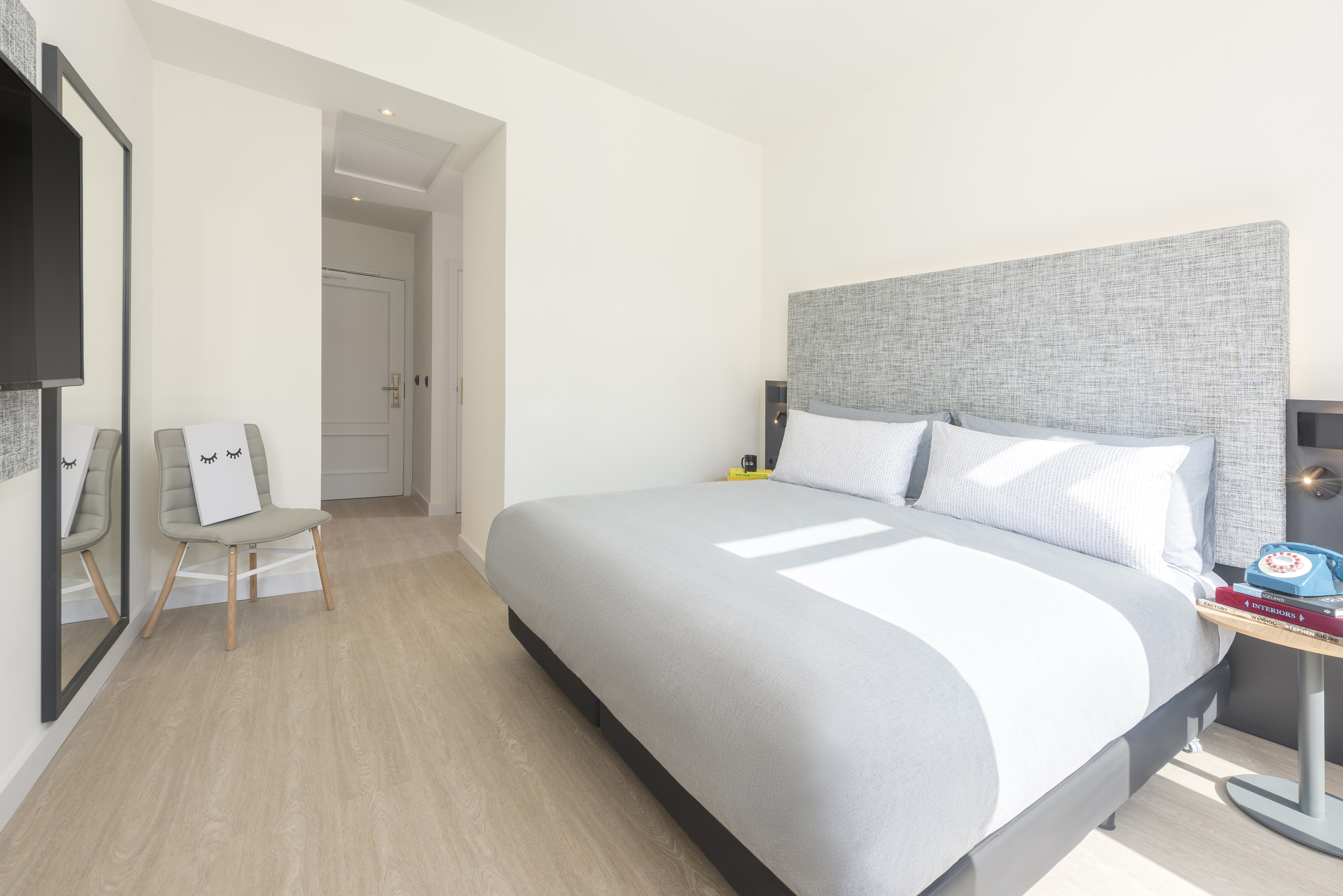 Modern hotel bedroom, grey bed, light wood floor, minimalist style.