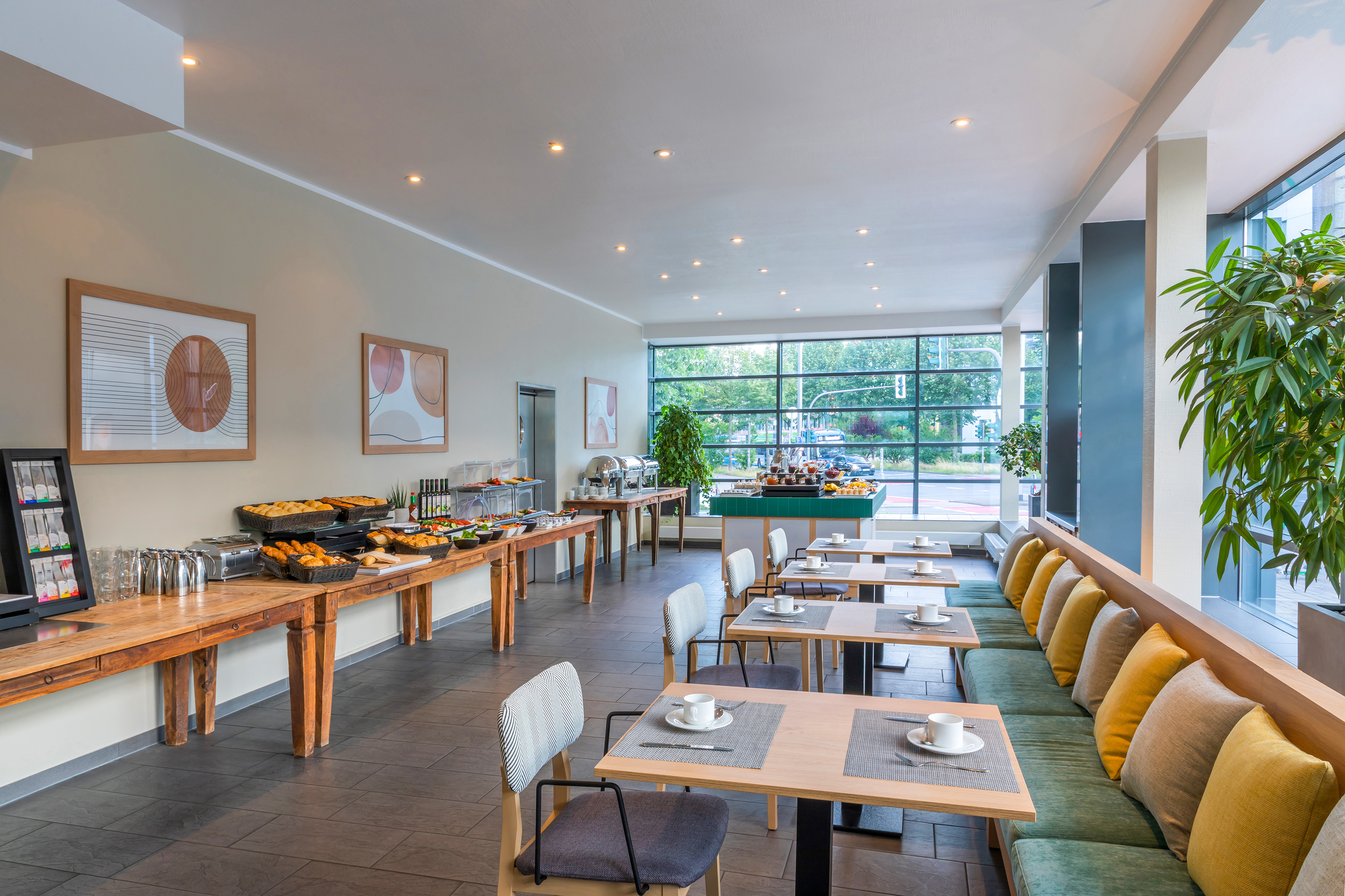 Hotel breakfast room with buffet, dining tables, and window seating area.