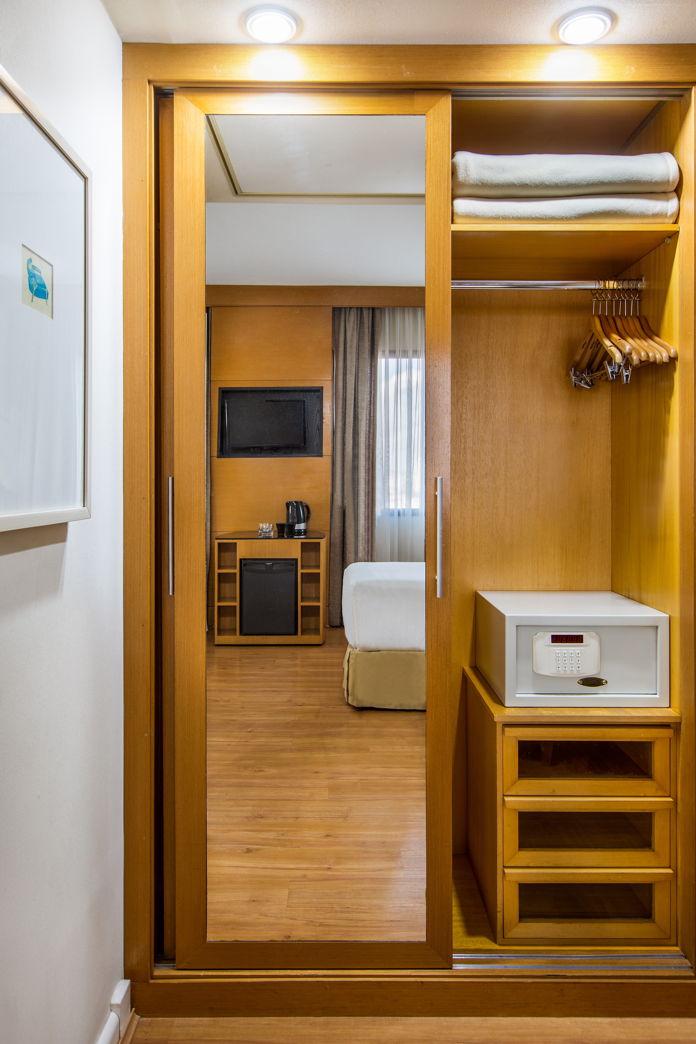 Hotel room closet with mirror, safe, towels, reflecting bed, TV, minibar.