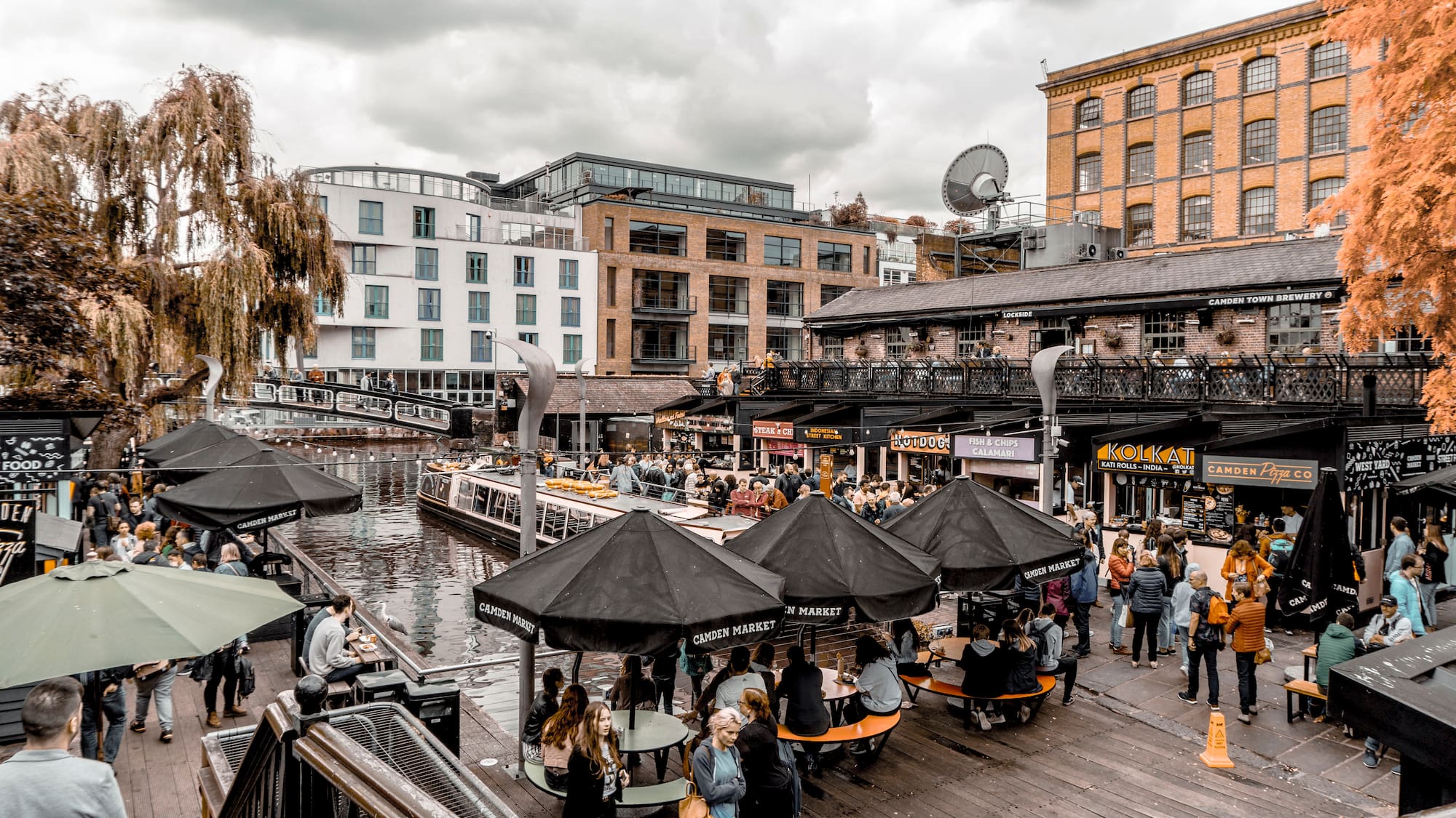 A group of people stands outside in a city, gathered under umbrellas and around tables.